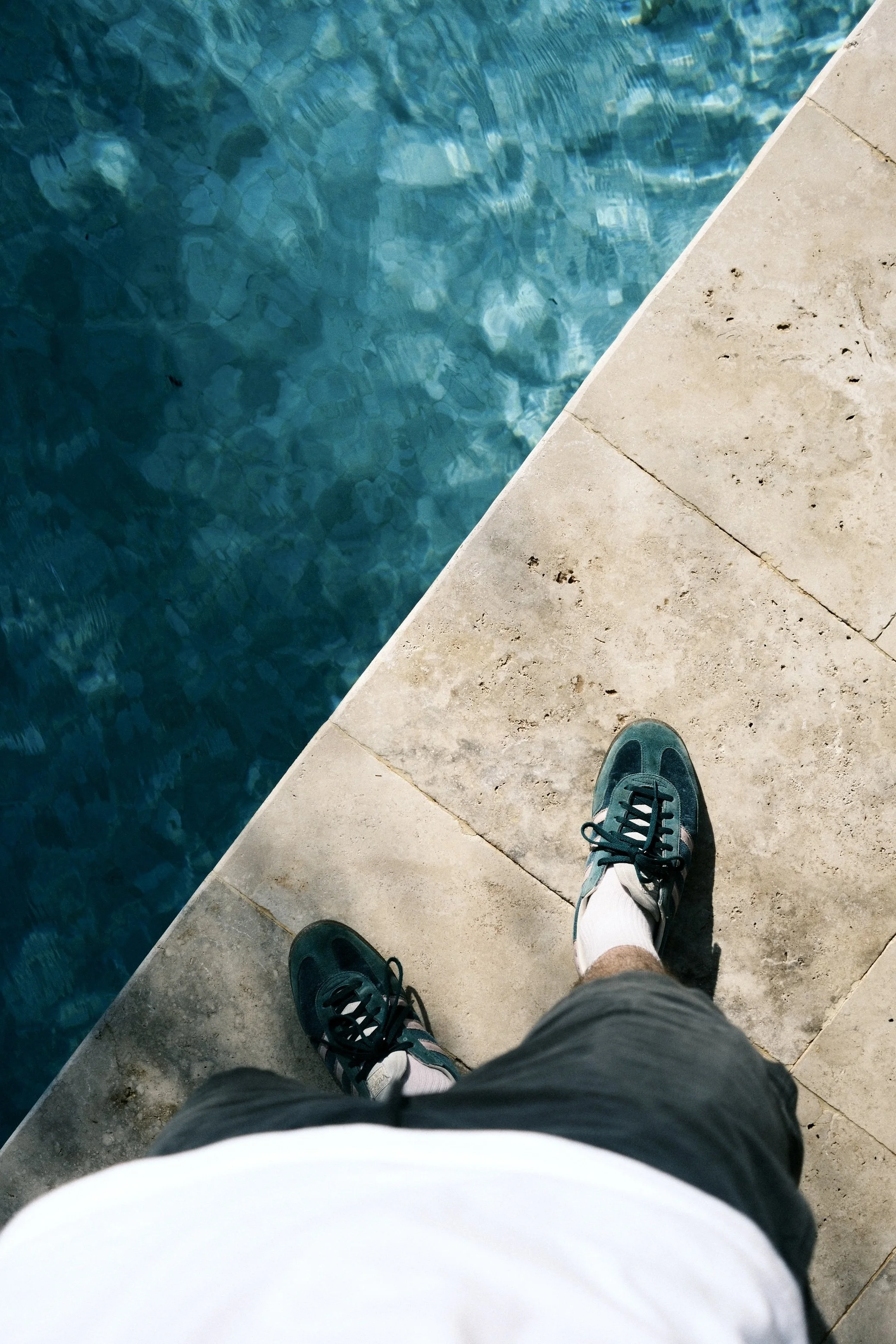 Prise de vue du regard d'une personne debout sur le bord d'une piscine, portant des chaussures de sport, des chaussettes blanches, des shorts et un t-shirt blanc, avec l'eau bleue visible à côté.
