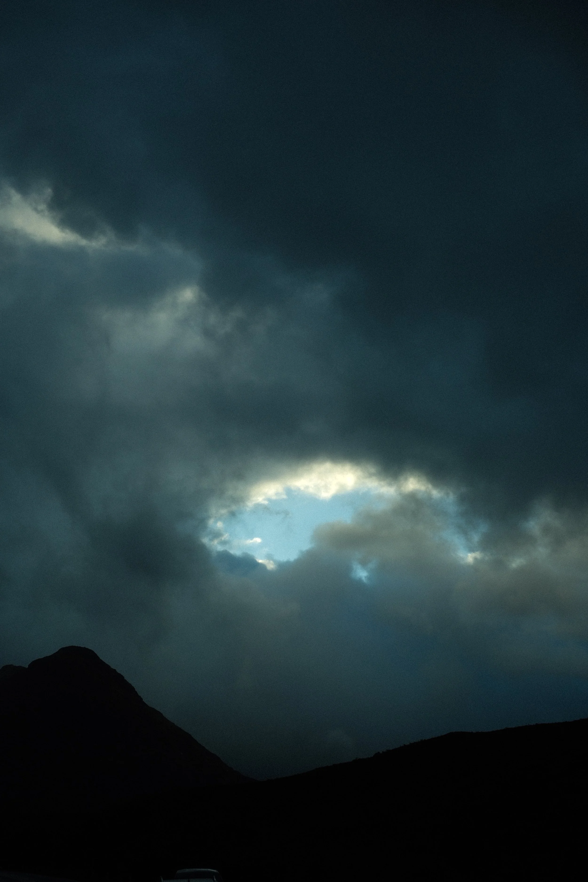Ciel nuageux avec une ouverture laissant entrevoir un peu de ciel bleu derrière des nuages sombres, silhouette d'une montagne ou d'une colline en bas.