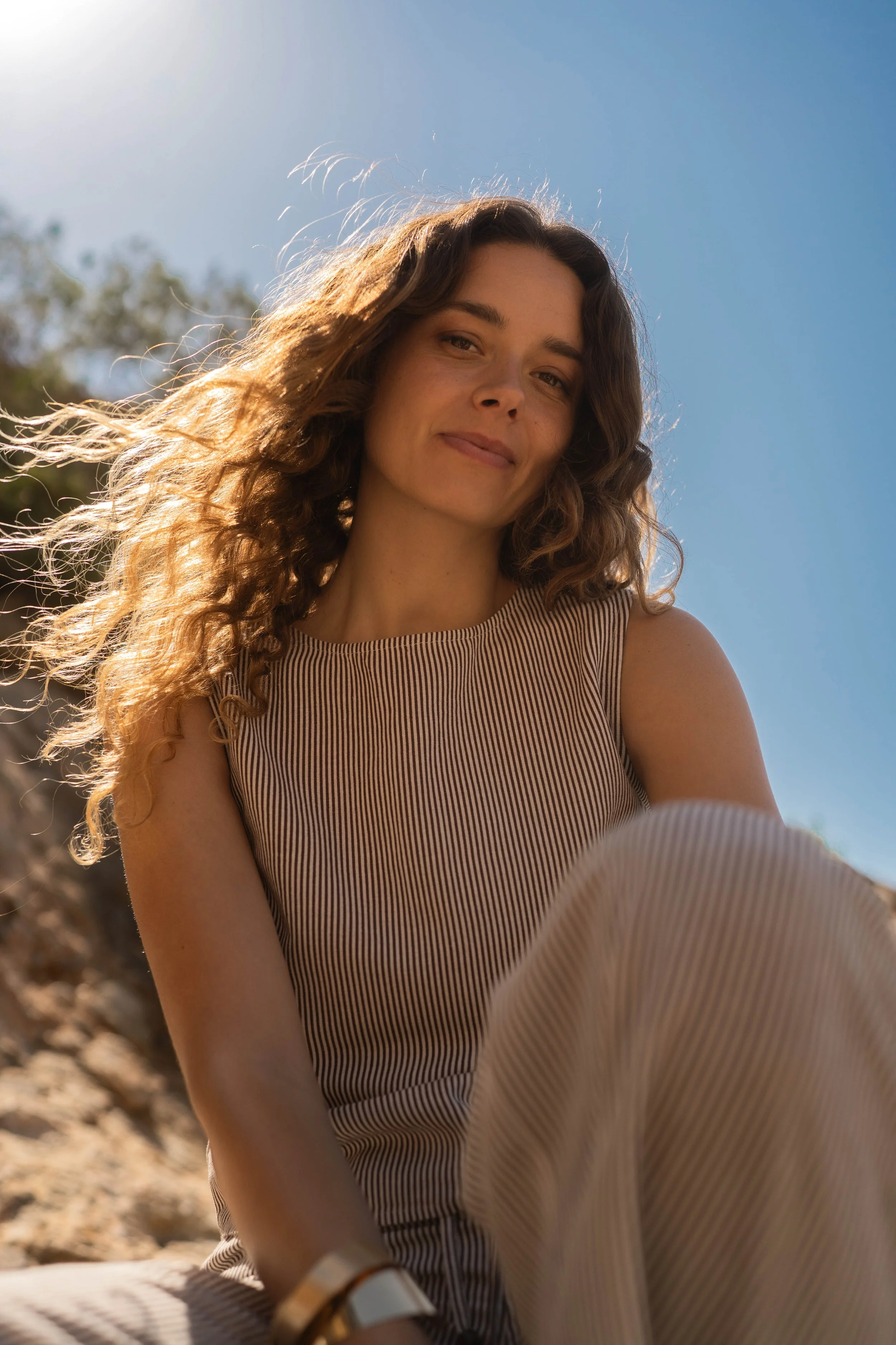 Une jeune femme aux cheveux bouclés et longs, portant une tenue à rayures, assise à l'extérieur sous un ciel bleu, avec le soleil qui brille derrière elle.