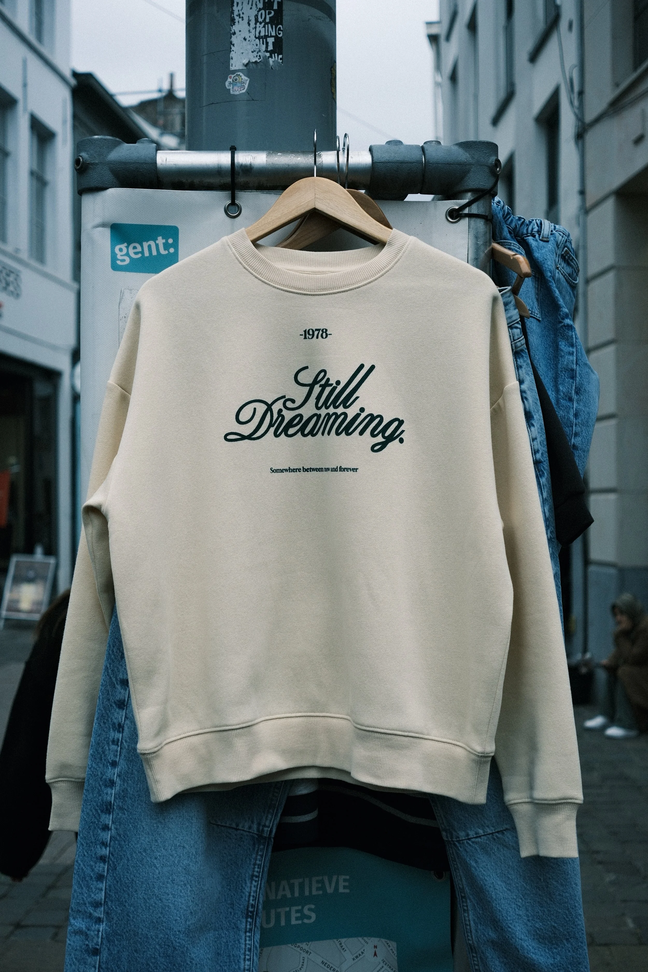 Un sweat-shirt beige avec l'inscription 'Still Dreaming' suspendu sur un support, à côté d'un pantalon en jean bleu, sur une rue urbaine