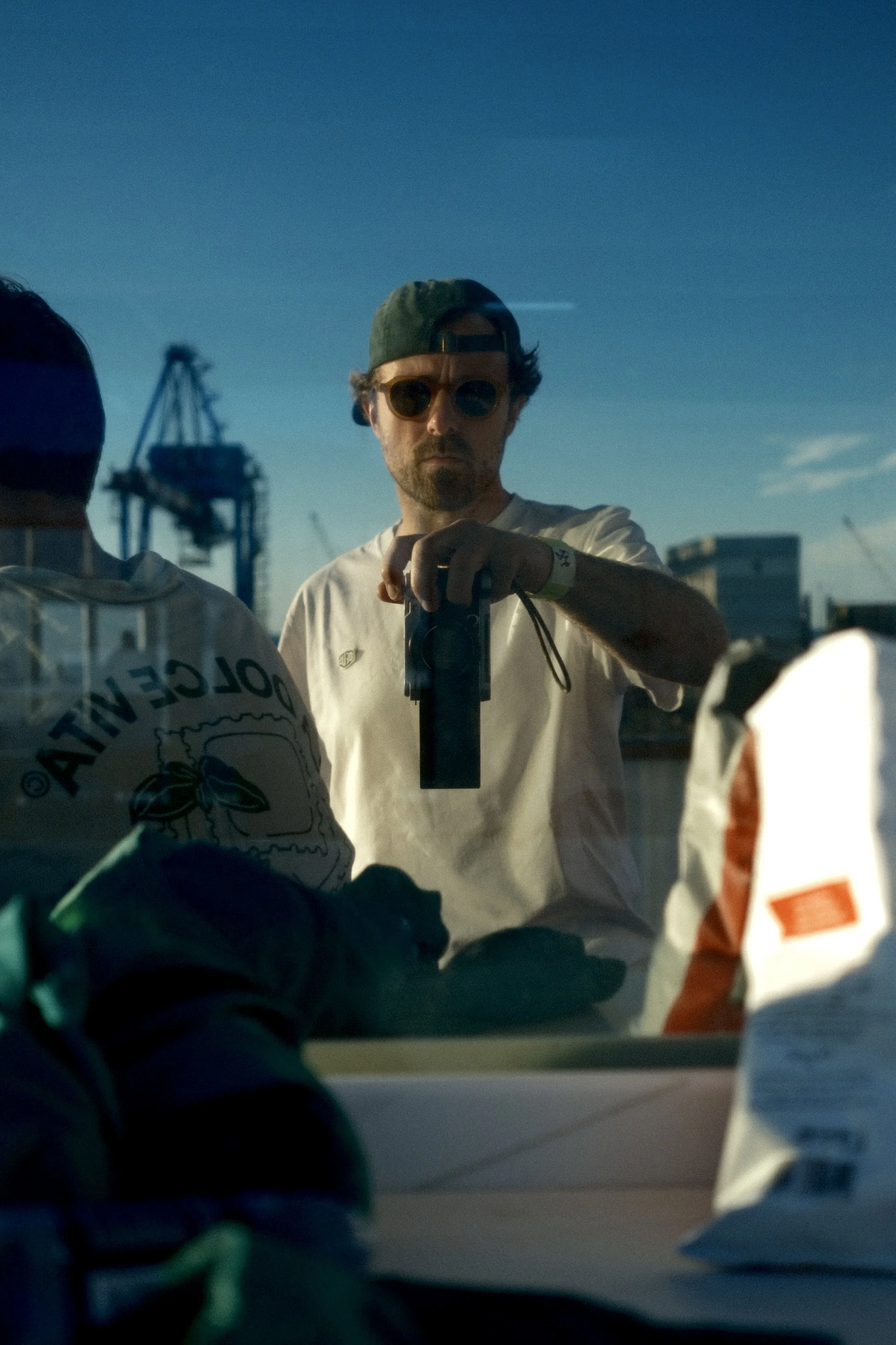 Un homme portant des lunettes de soleil et un t-shirt blanc, en train de prendre une photo ou de se filmer devant une fenêtre, avec une vue du ciel bleu et des bâtiments en arrière-plan.