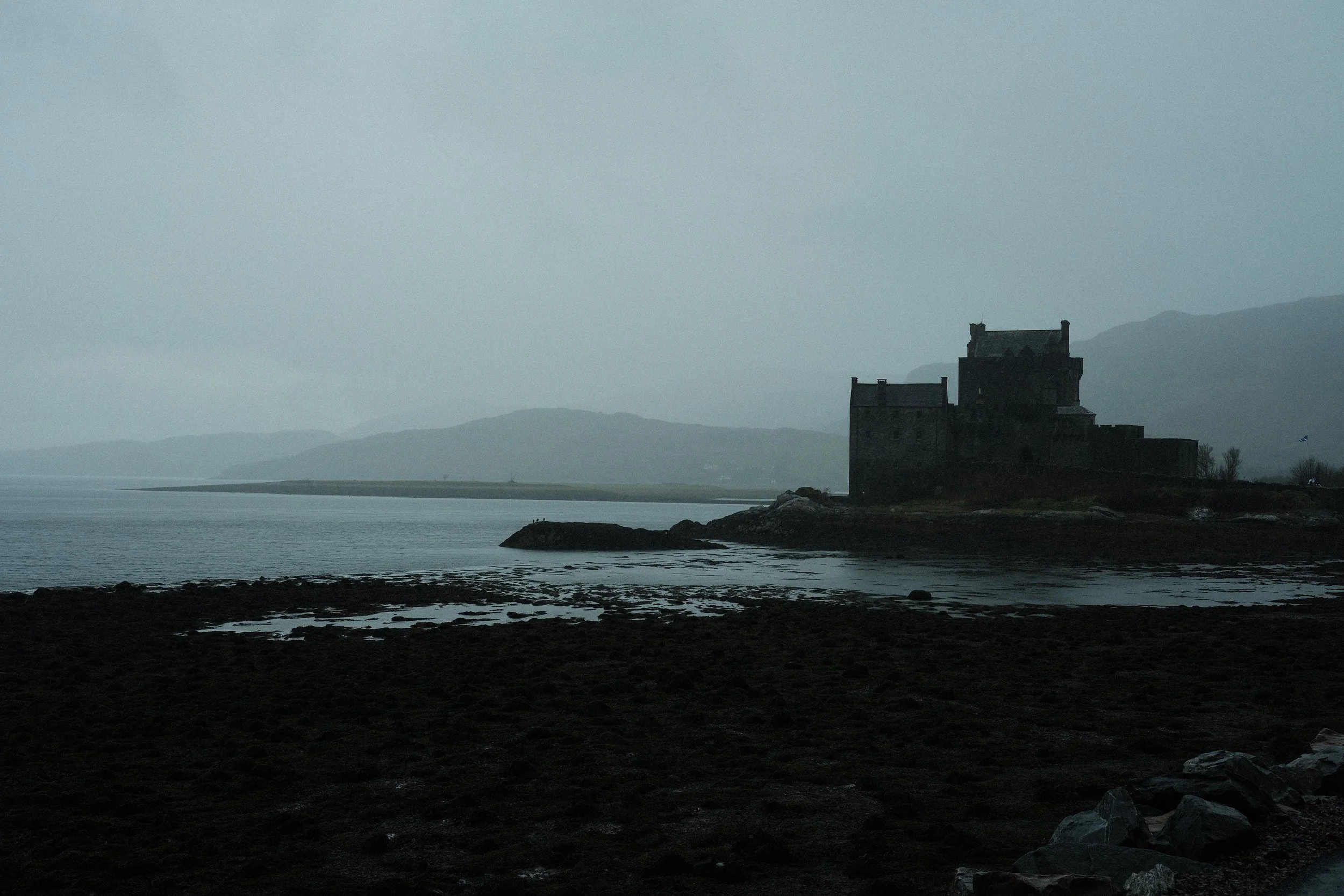 Château ancien en pierre sur une petite île au bord de l'eau dans un paysage brumeux et montagneux.