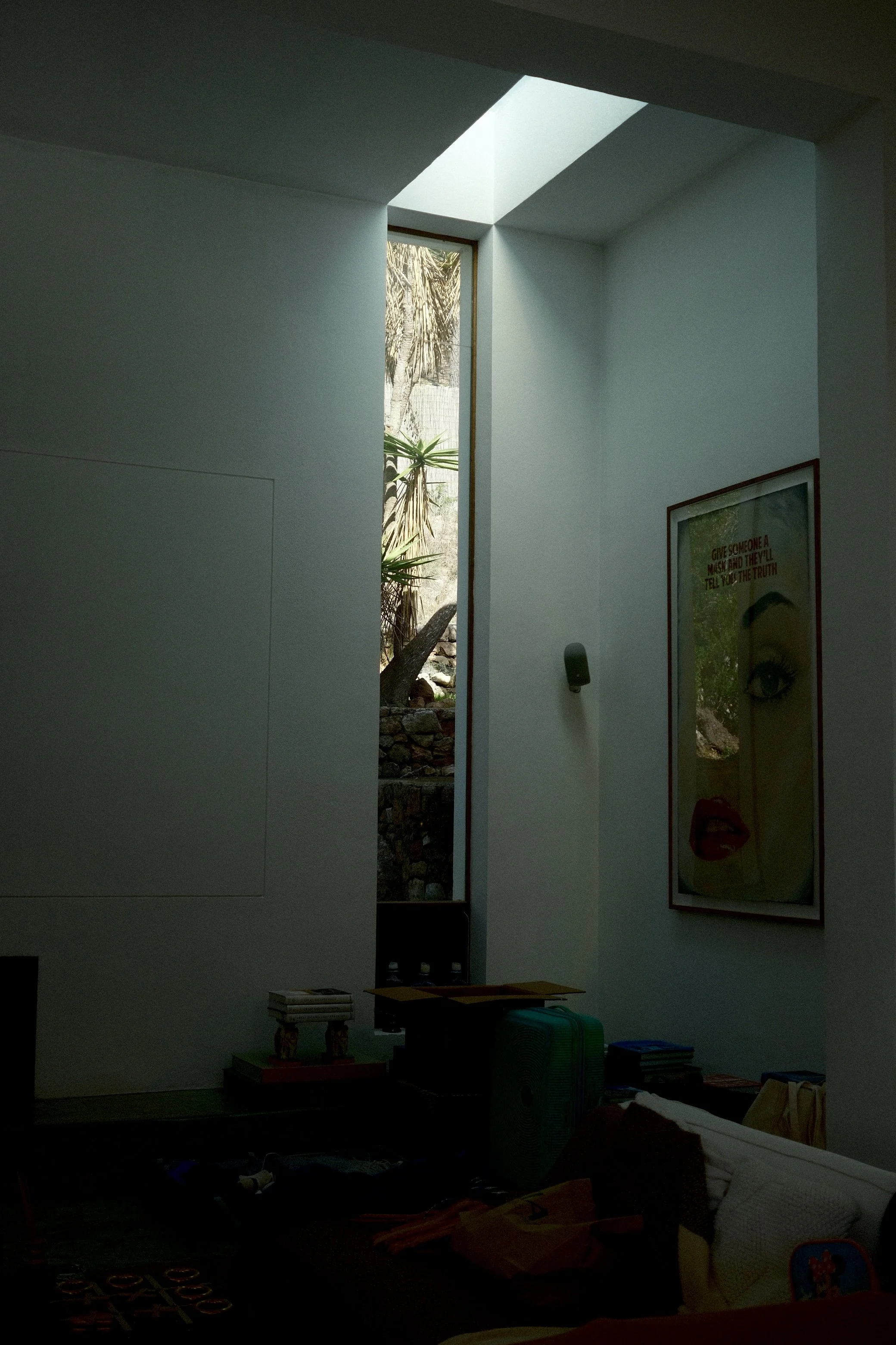 Une pièce avec un mur blanc, une fenêtre étroite laissant entrer de la lumière naturelle, un tableau avec un visage et un texte, et divers objets sur une surface en bas.