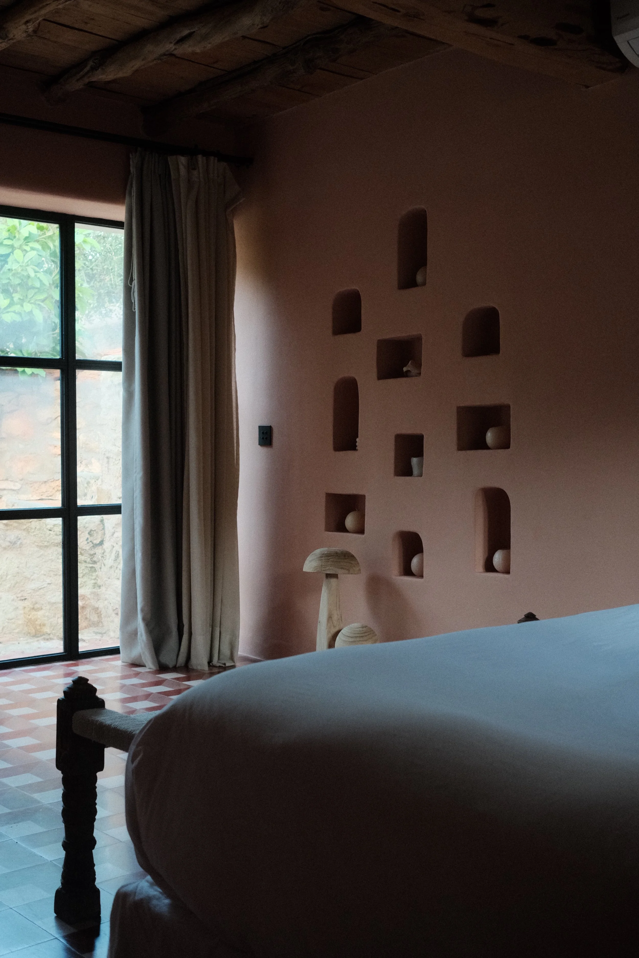 Chambre avec mur rose avec niches decoratives, grand lit avec draps blancs, porte-fenêtre avec rideaux blancs, sol en carreaux, lampe en bois près du lit.