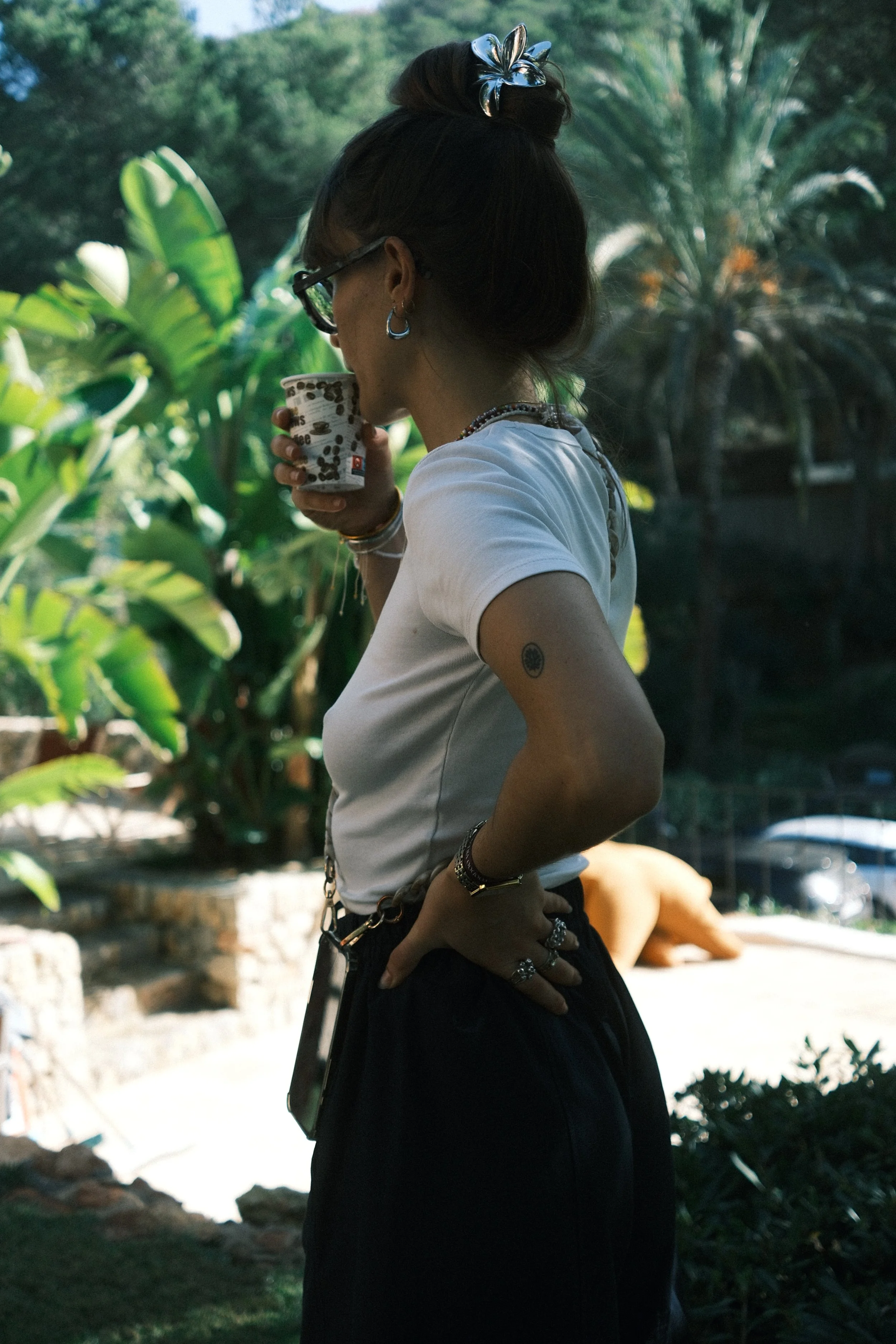 Femme portant des lunettes et des bijoux, tenant une tasse à café, debout à l'extérieur près de plantes vertes et d'arbres.