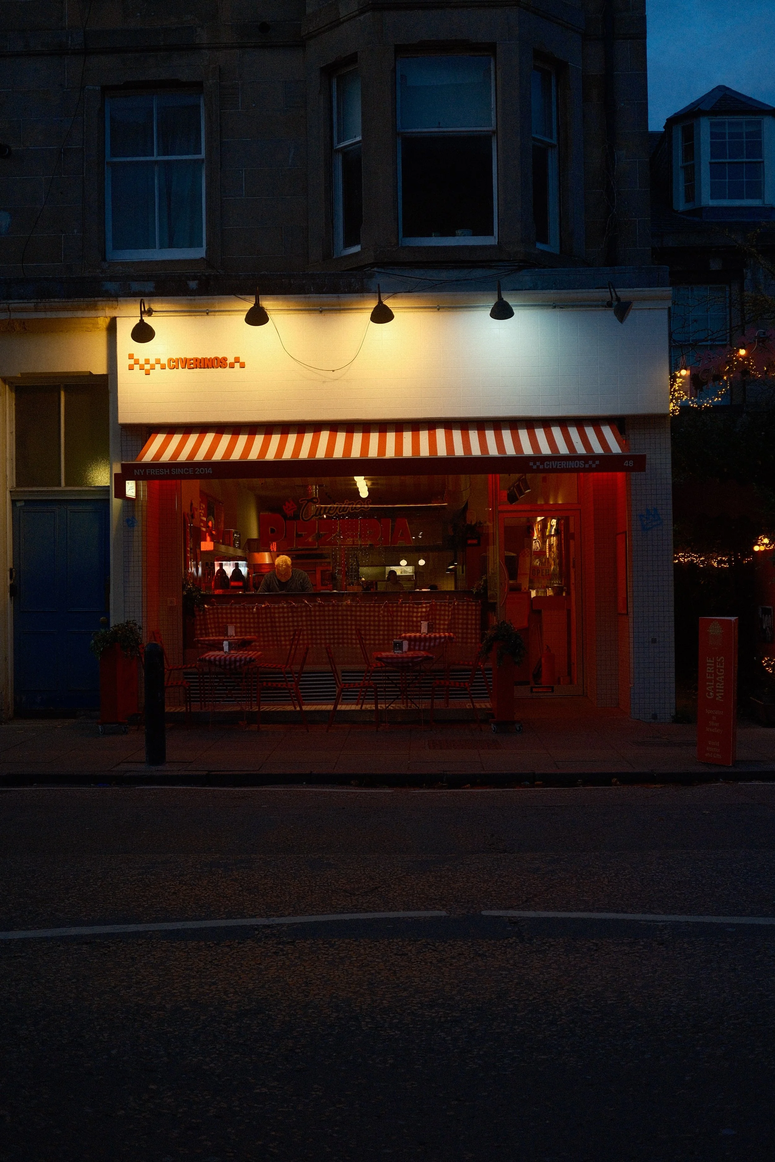 Une pizzeria ouverte la nuit avec des tables dehors, un éclairage chaud et un homme derrière le comptoir. Façade avec enseignes en rouge et blanc, et un balcon au-dessus.