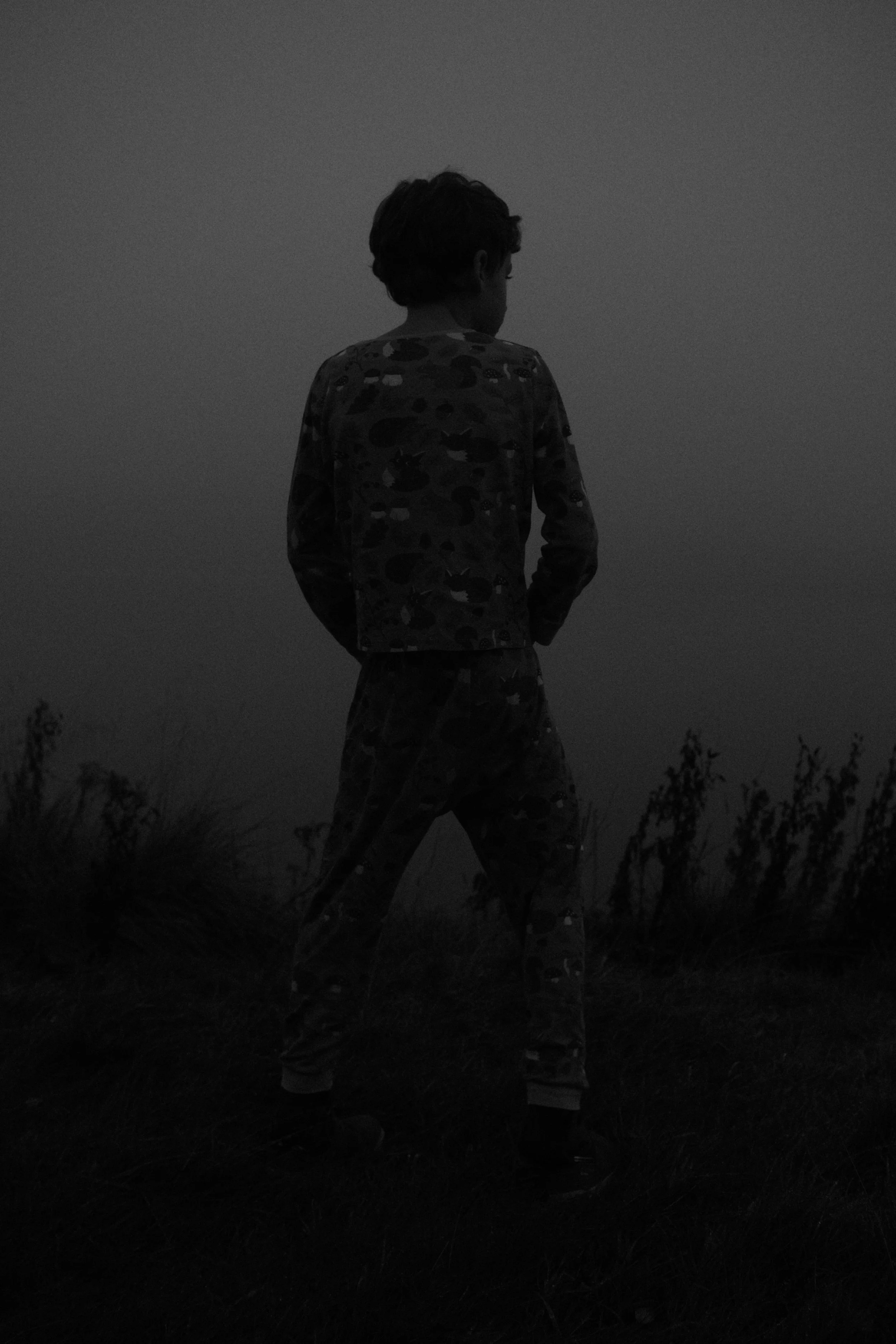 Un garçon vêtu d'un pyjama à motifs camouflages se tient dans un champ, vu de dos, dans une ambiance sombre ou en noir et blanc.