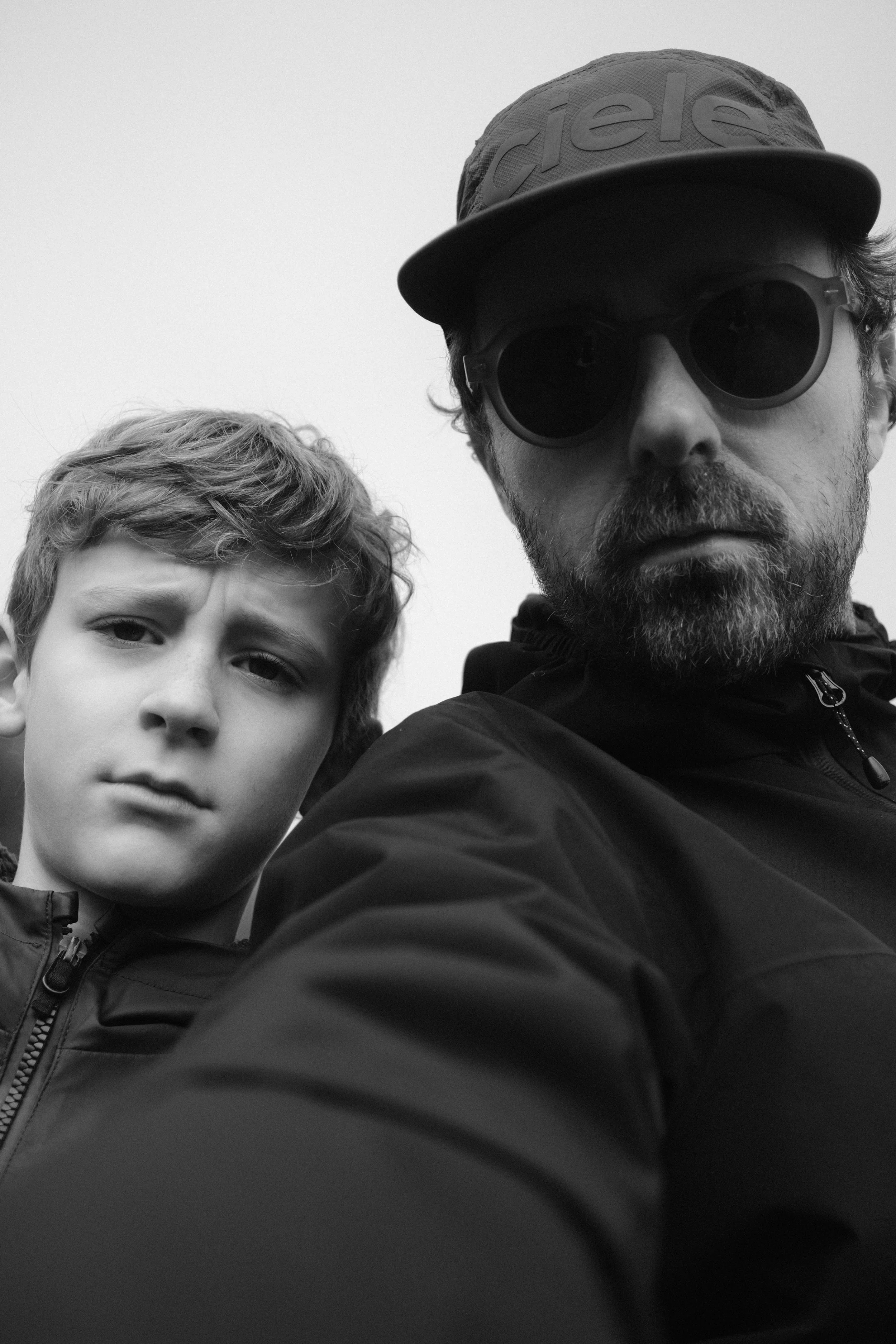 Un homme portant des lunettes de soleil, une casquette et une veste, à côté d'un garçon avec une expression sérieuse, tous deux en noir et blanc.