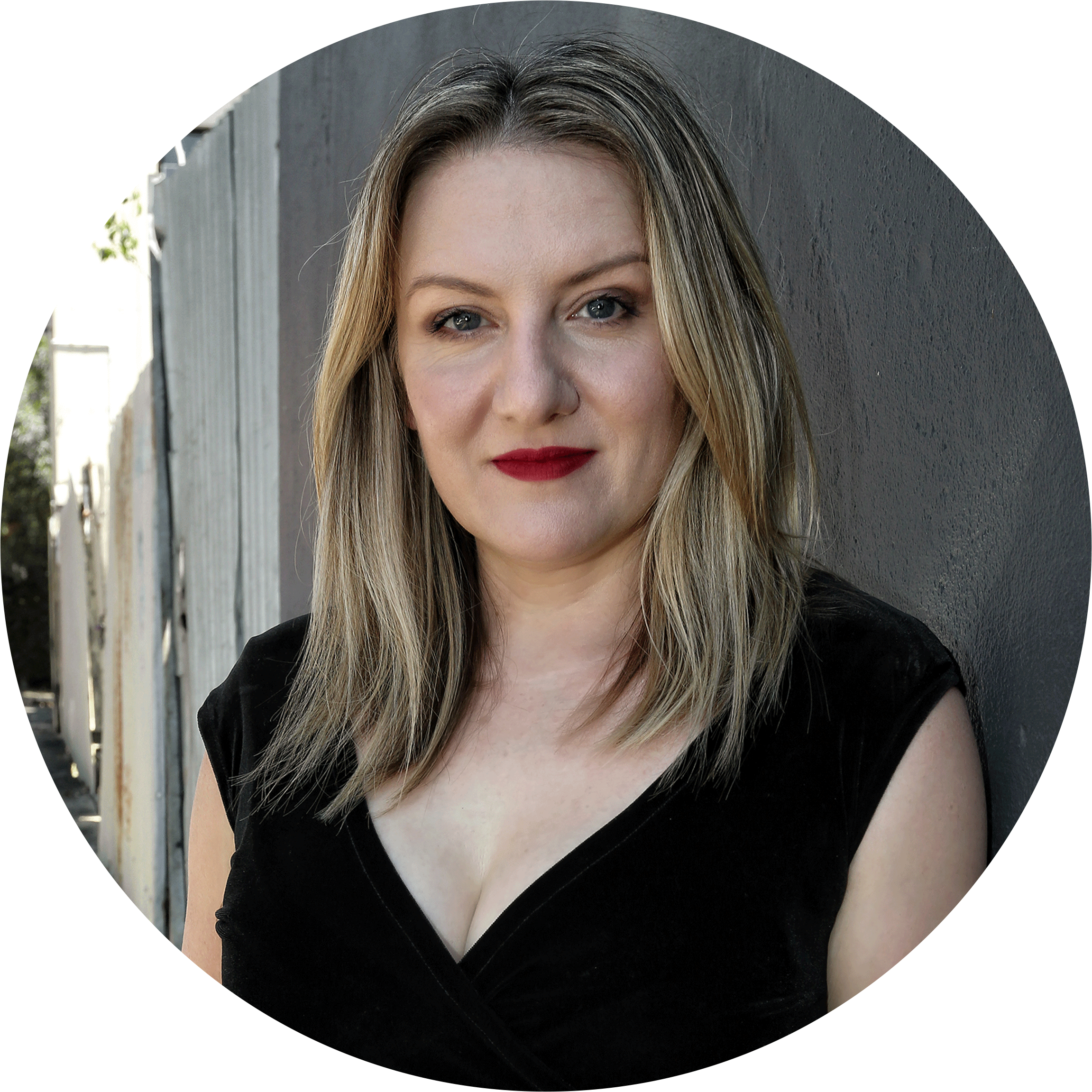 A woman with blonde hair, blue eyes, and red lipstick standing outdoors against a gray wall, wearing a black sleeveless top.