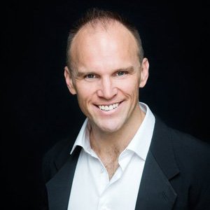 Portrait of a smiling man with a receding hairline, wearing a black blazer and white shirt, against a dark background.