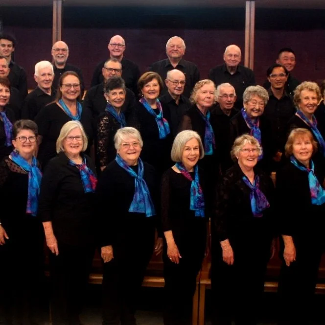 Group of choir members, mostly women, wearing black outfits with blue scarves, posing on stage.