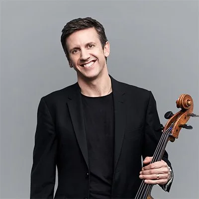 A smiling man in a black blazer holding a violin against a plain gray background.