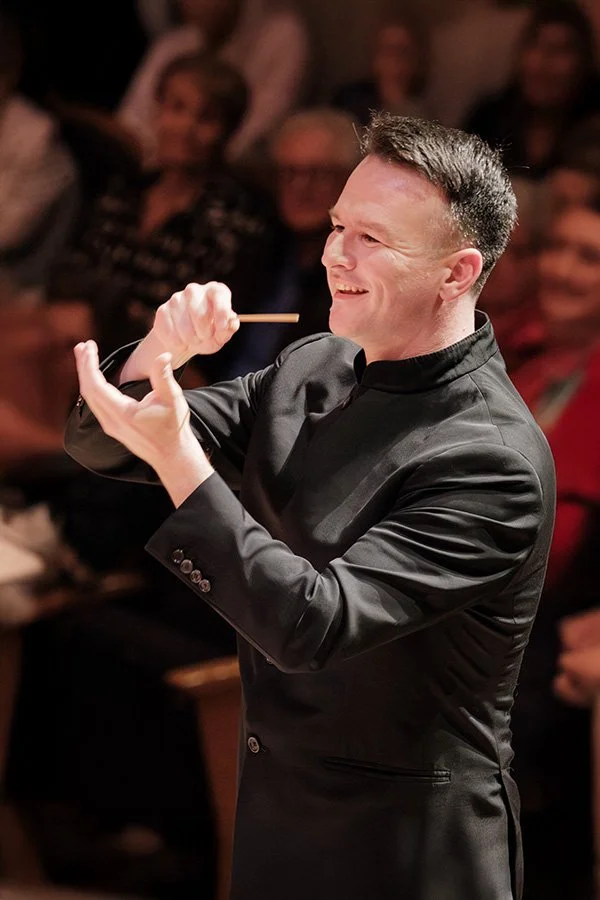 Conductor leading an orchestra, smiling, with an audience in the background.