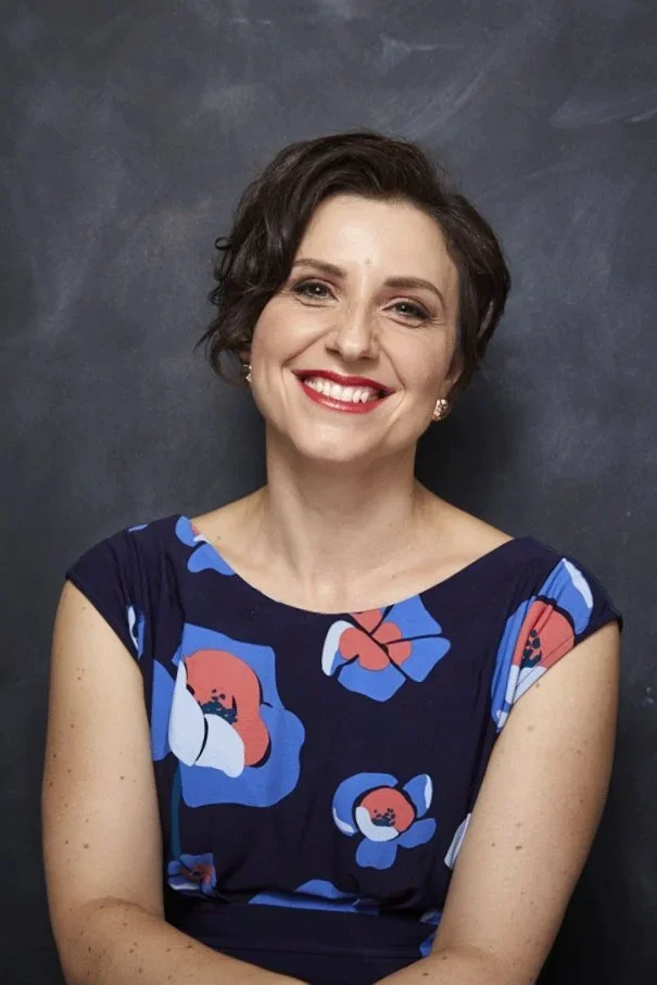 A woman with short wavy dark hair smiling, wearing a blue dress with large floral prints, standing against a dark background.