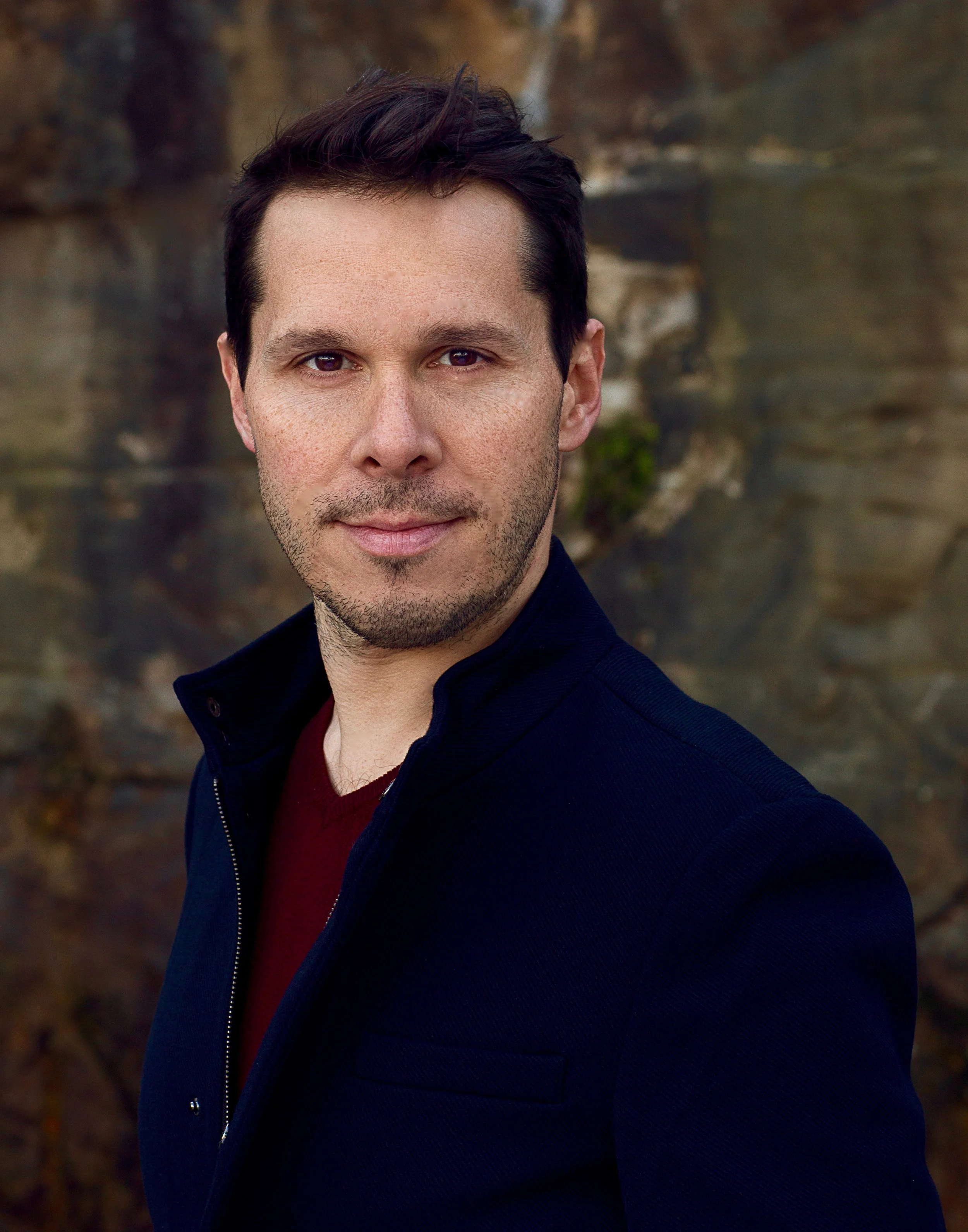 Portrait of a man with short dark hair, wearing a dark jacket over a red shirt, standing outdoors with rocks in the background.