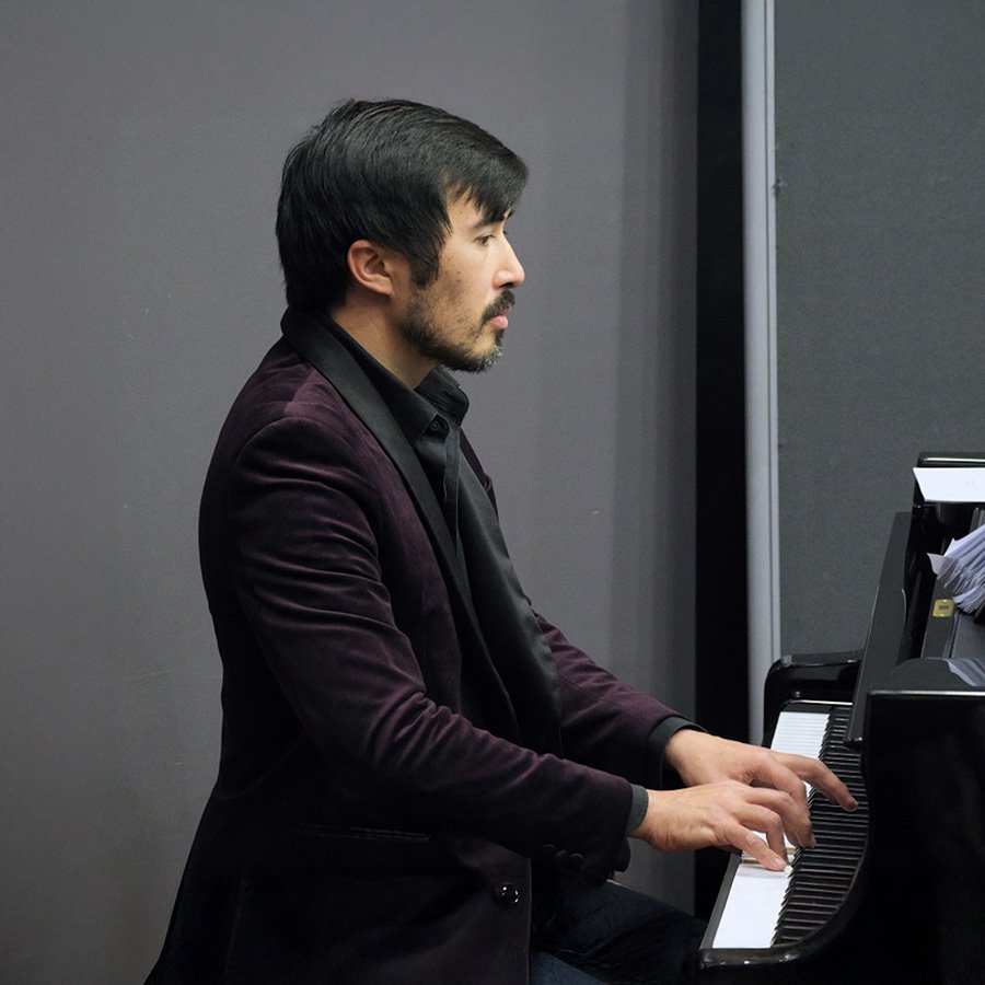 A man with dark hair and a beard playing a black upright piano in a room with dark gray walls.