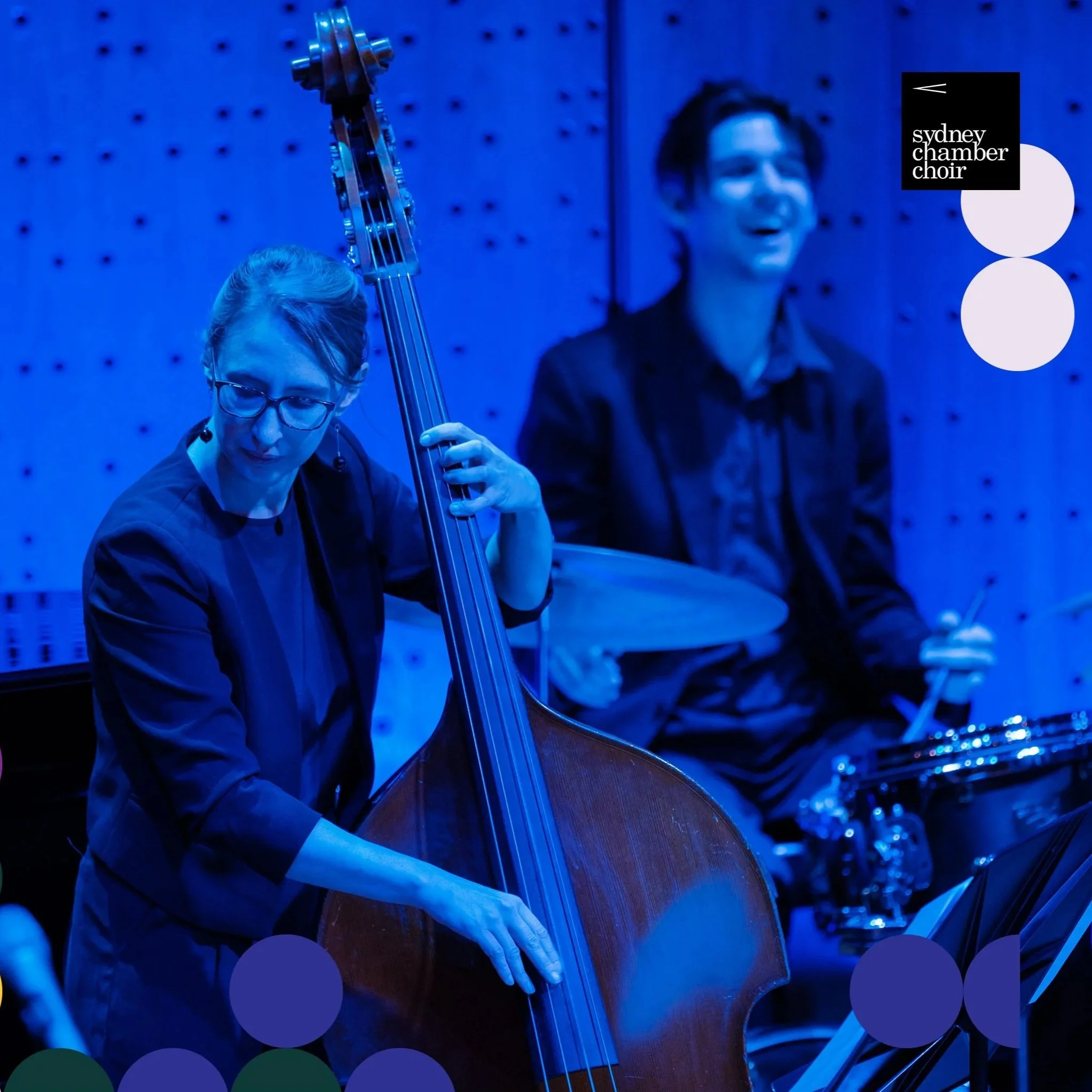 Musicians performing at Sydney Chamber Choir concert, with a woman playing upright bass and a man playing drums, in a blue-lit room.