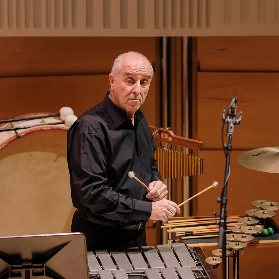 An older man in a black shirt holding mallets stands behind a percussion instrument setup, including a xylophone, cymbals, and drums, in a wooden-paneled music studio or concert hall.