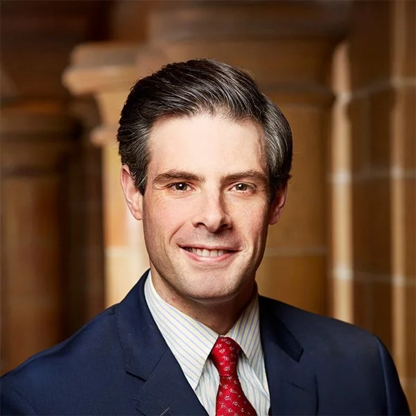 Smiling man in a navy suit, light blue striped shirt, and red tie posing in a wooden setting.