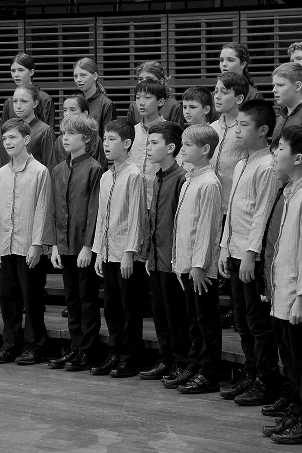 A group of children singing in a choir on stage, arranged in rows, dressed in coordinated outfits.