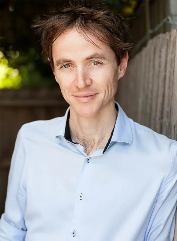 A young man with brown hair, wearing a light blue button-up shirt, smiling outdoors near a wooden fence with greenery in the background.