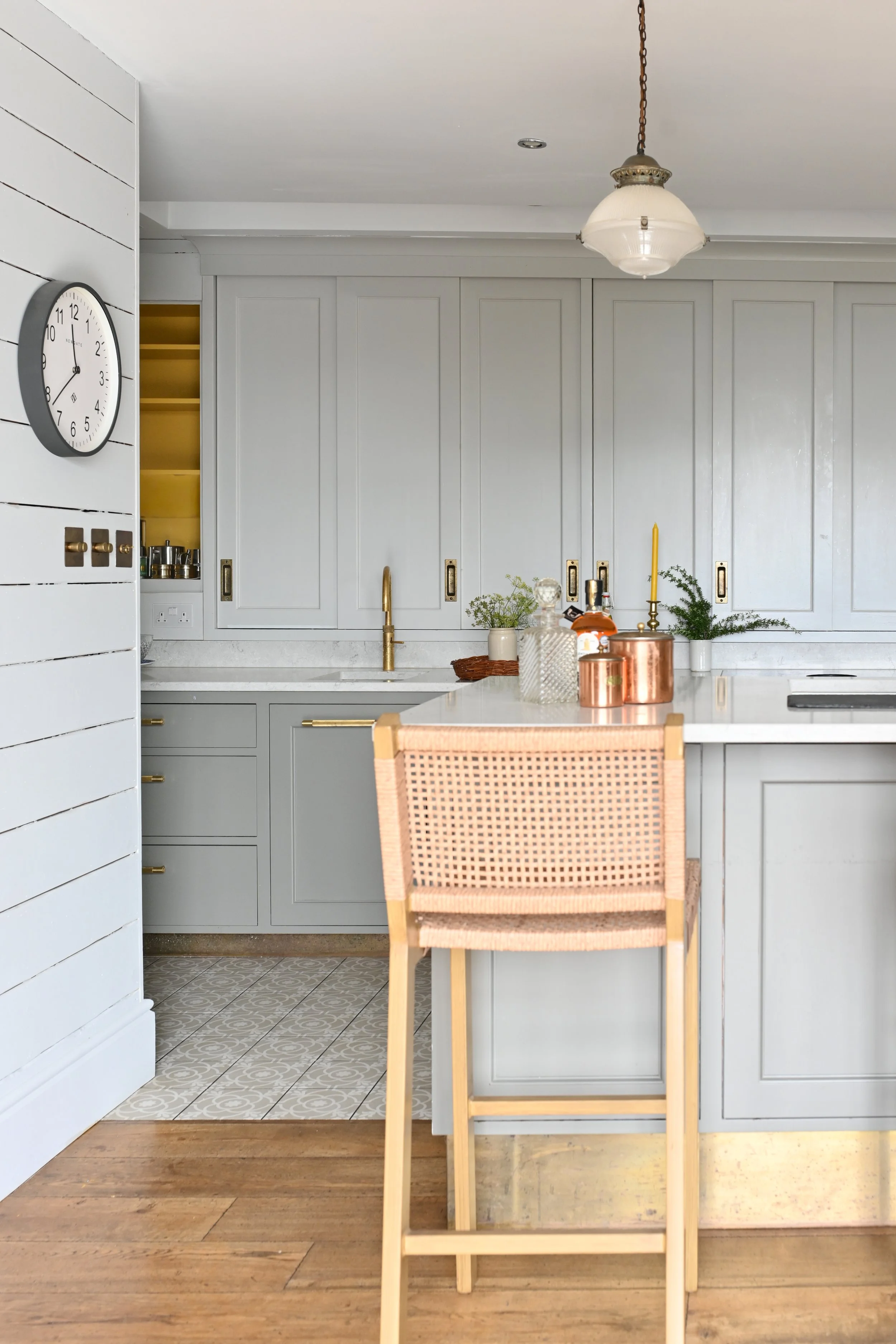 A cozy kitchen with light gray cabinets, a gold faucet, and a white marble countertop. A round clock on the wall shows 9:55. Modern pendant lighting hangs from the ceiling. There are small plants and decorative items on the counter, and a woven ratta