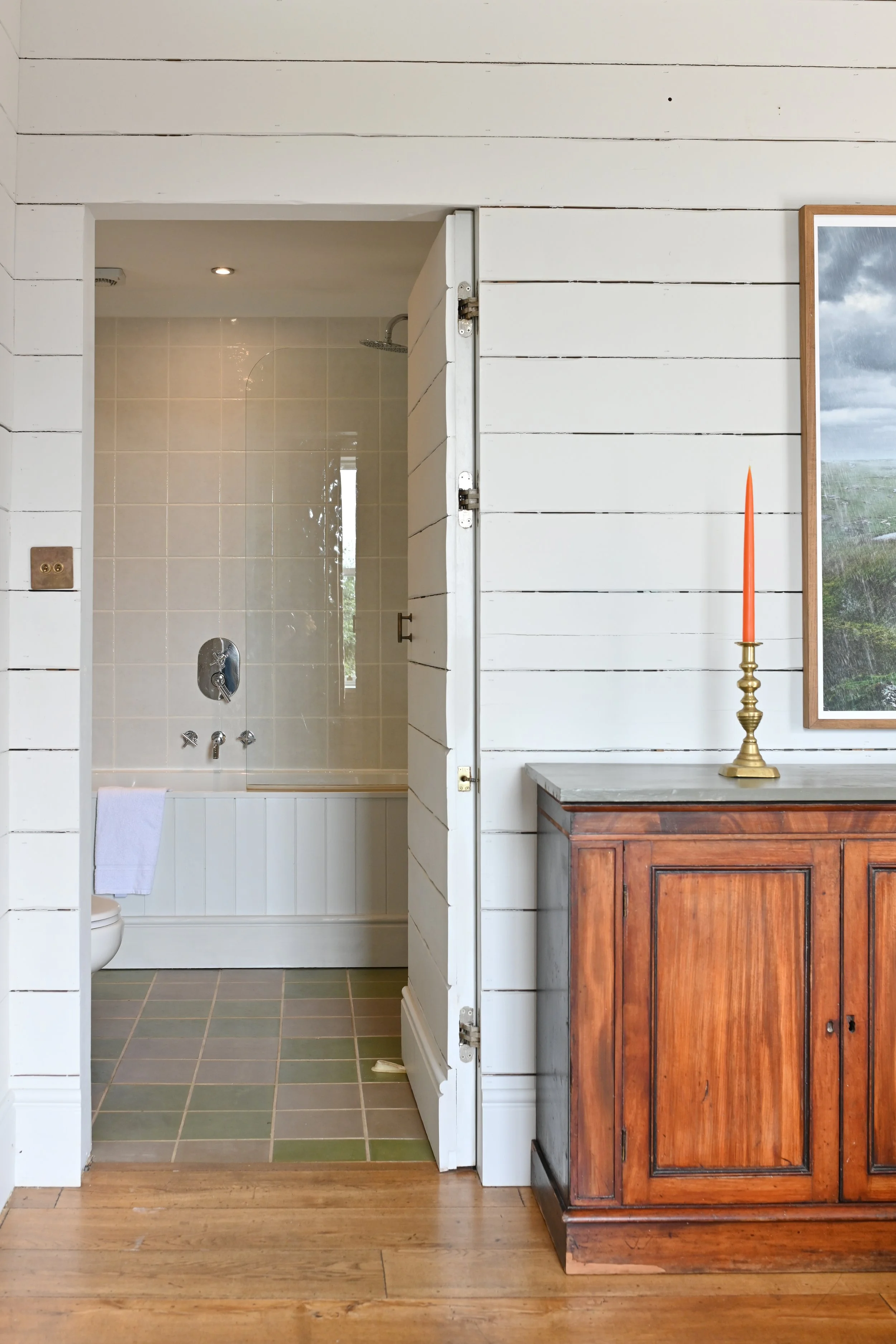 Interior of a house showing a bathroom with a bathtub and a shower, and a wooden cabinet with a candle on top, next to a framed landscape photograph.