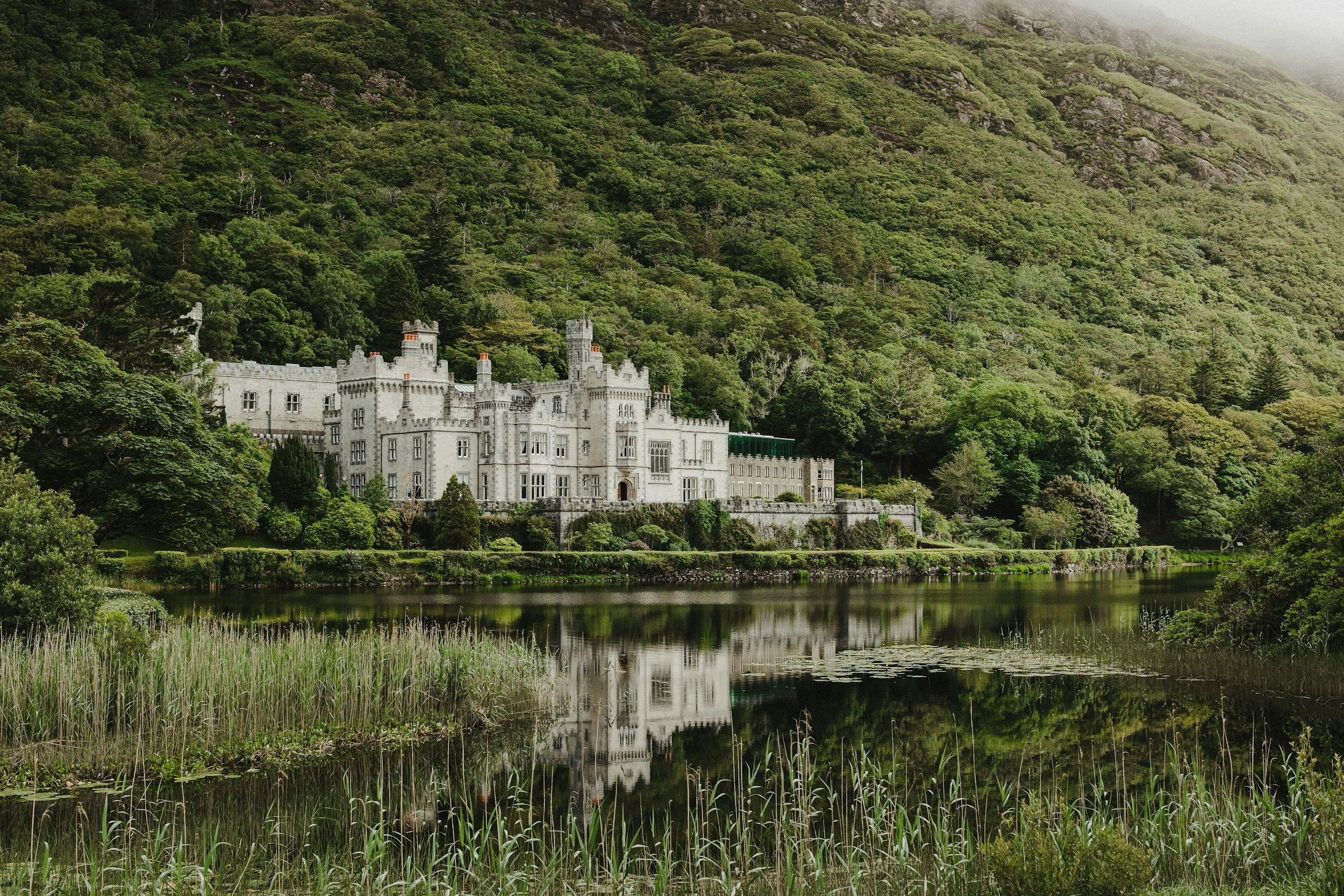 A large, white castle-style mansion with turrets and crenellated walls, situated by a calm water body, with lush green trees and a hillside in the background.