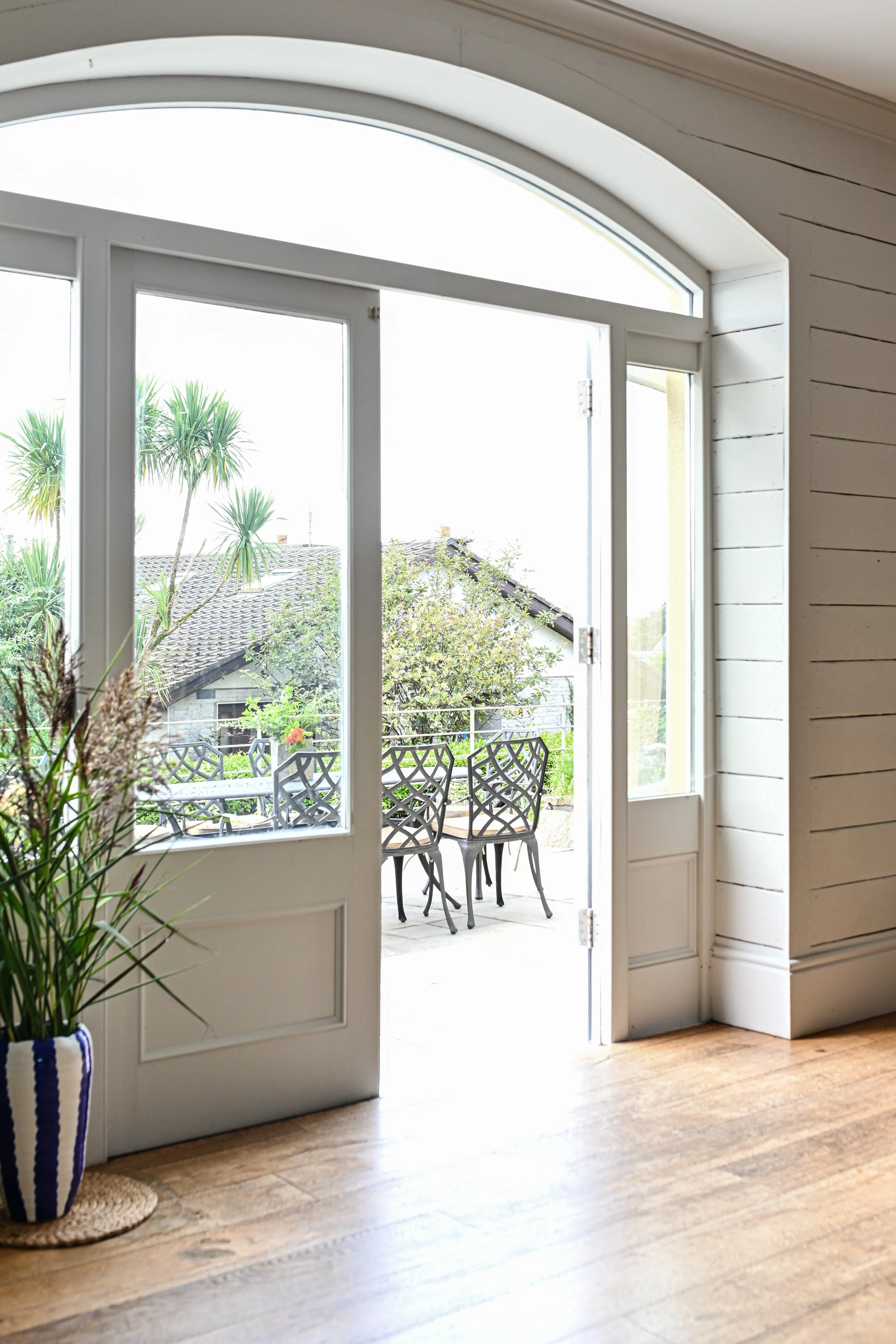 Open patio door revealing an outdoor seating area with metal chairs, surrounded by trees and greenery.