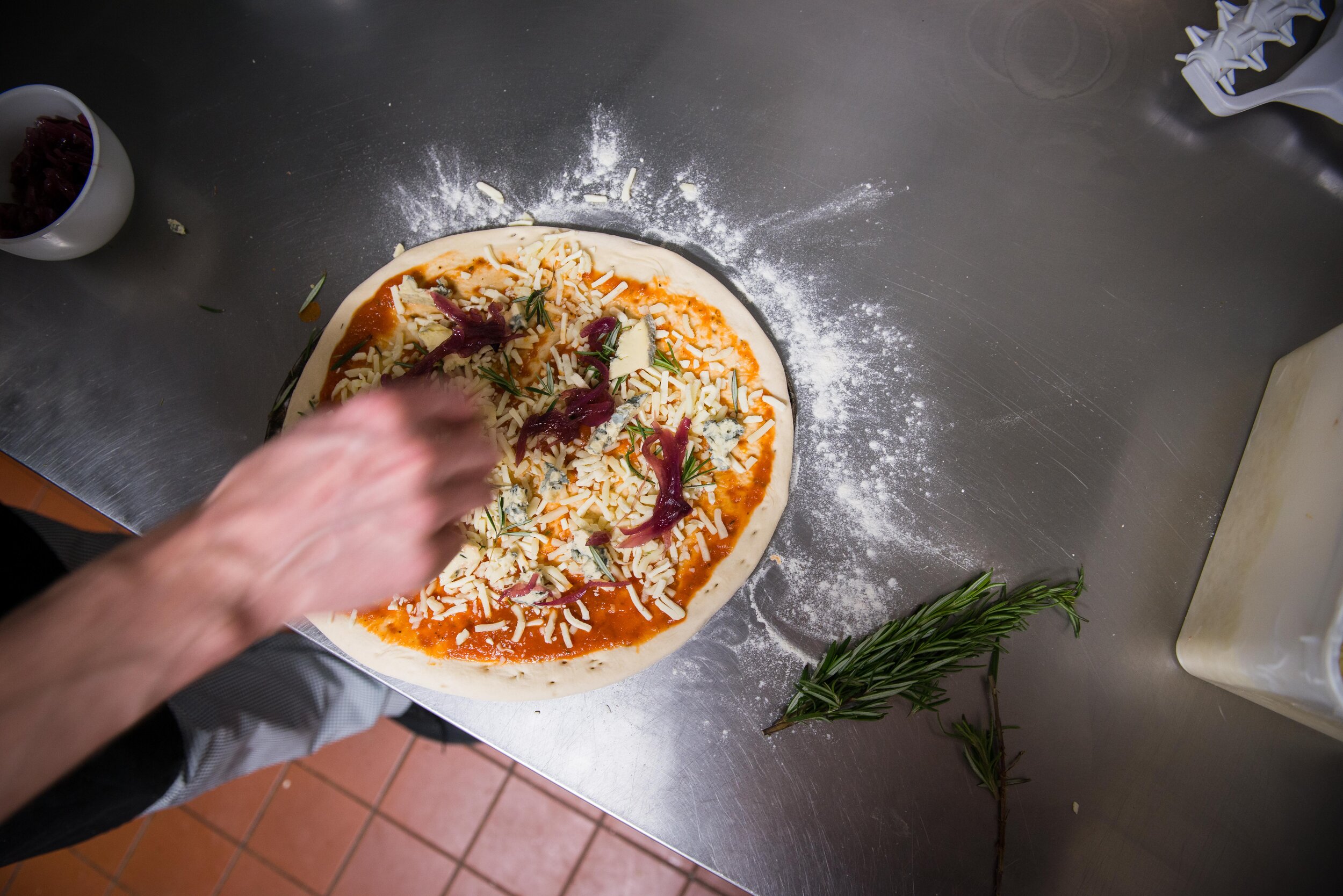 Person preparing a pizza on a floured countertop with tomato sauce, cheese, and toppings; a sprig of rosemary nearby.