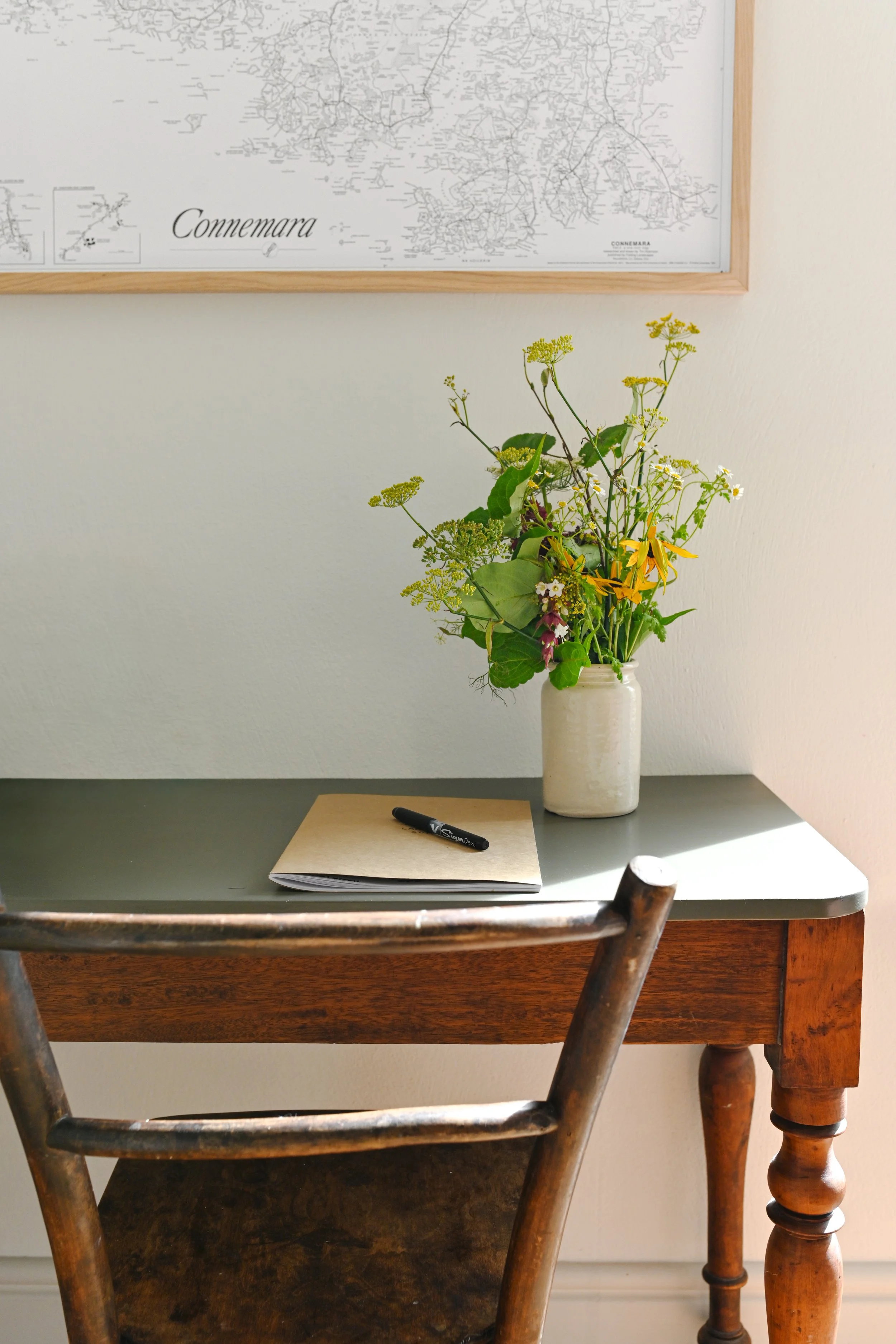 A wooden desk with a black top surface, a white vase with yellow and purple flowers, a closed notebook with a black pen on top, and a wooden chair in front of it, with a framed map on the wall behind.