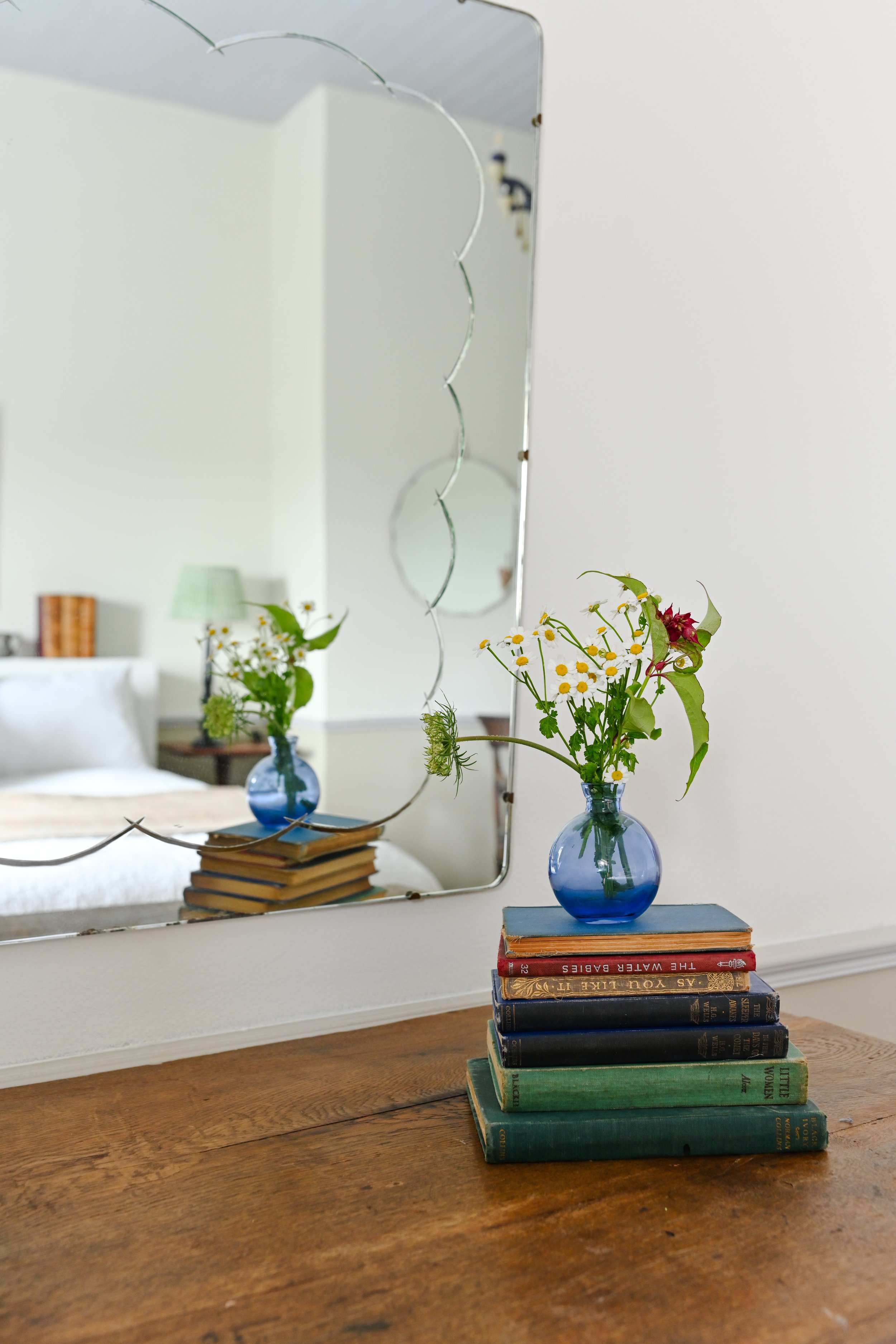A wooden table with a stack of vintage books and a blue glass vase holding wildflowers, reflected in a mirror on the wall.