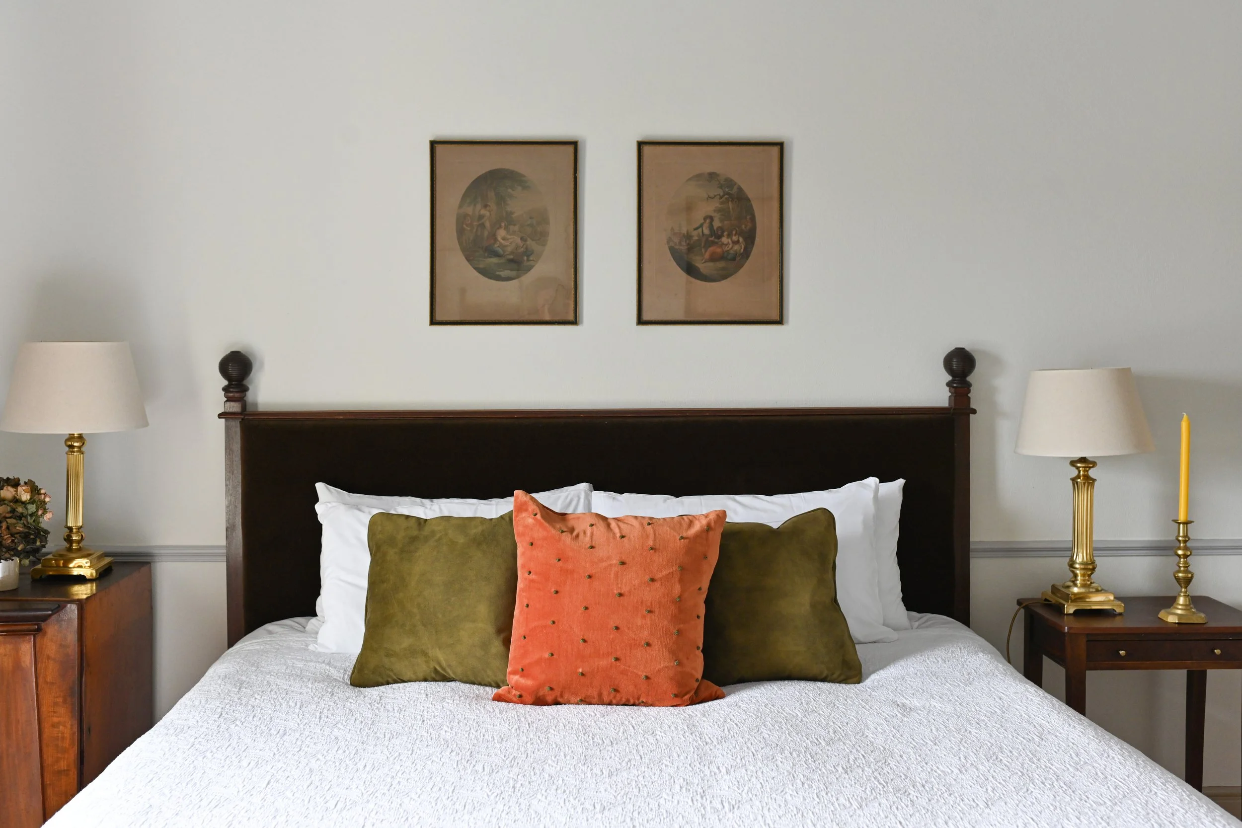 A bedroom with a bed that has a dark wooden headboard, three pillows (one orange with small black dots and two olive green), and white bedding. There are two bedside tables with lamps, and two framed vintage artwork prints hang above the bed.