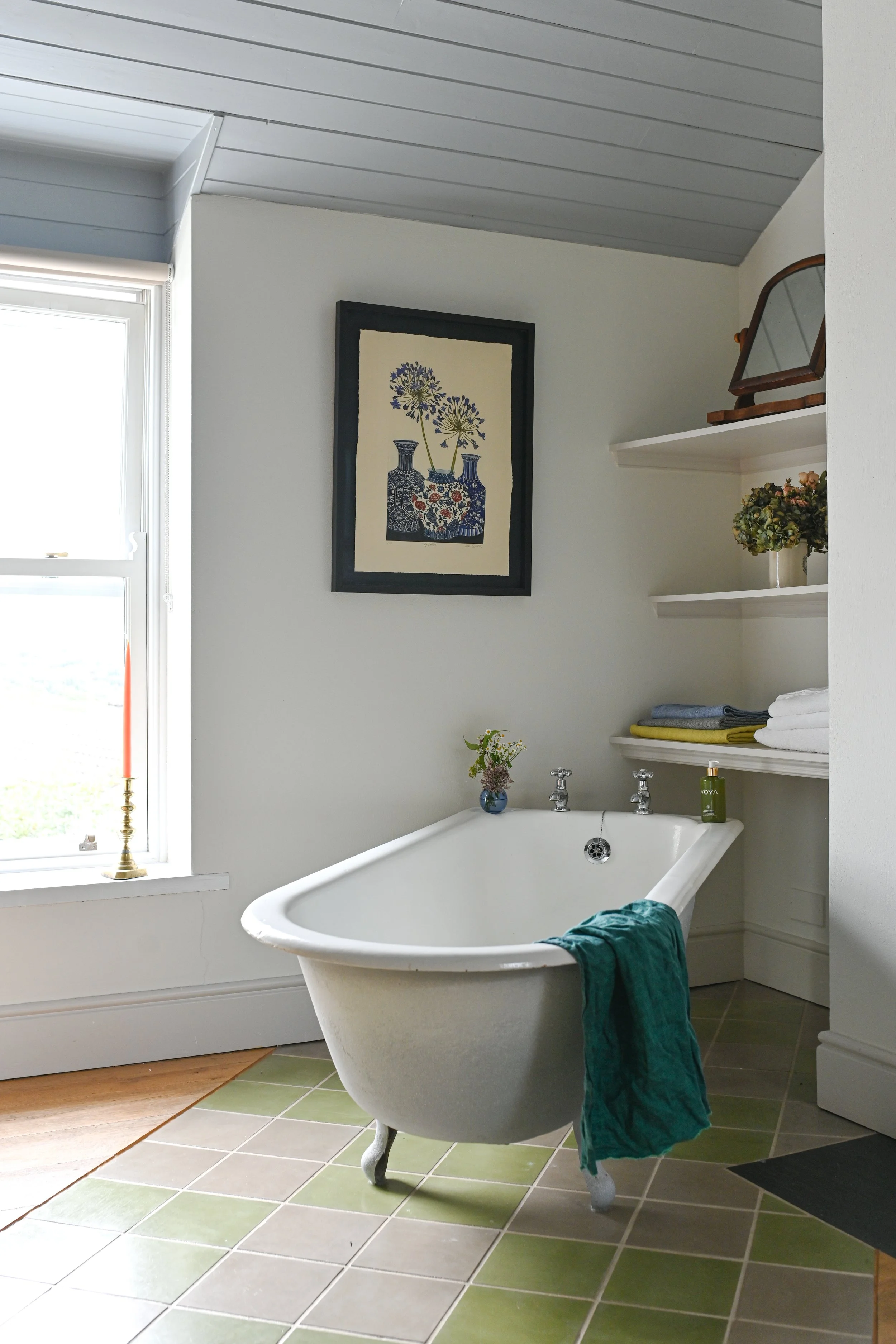 A vintage white clawfoot bathtub with a green towel hanging over the side, positioned in a bright bathroom with white walls, green tiled floor, a large window with sunlight streaming in, and shelves with towels and decorative items.