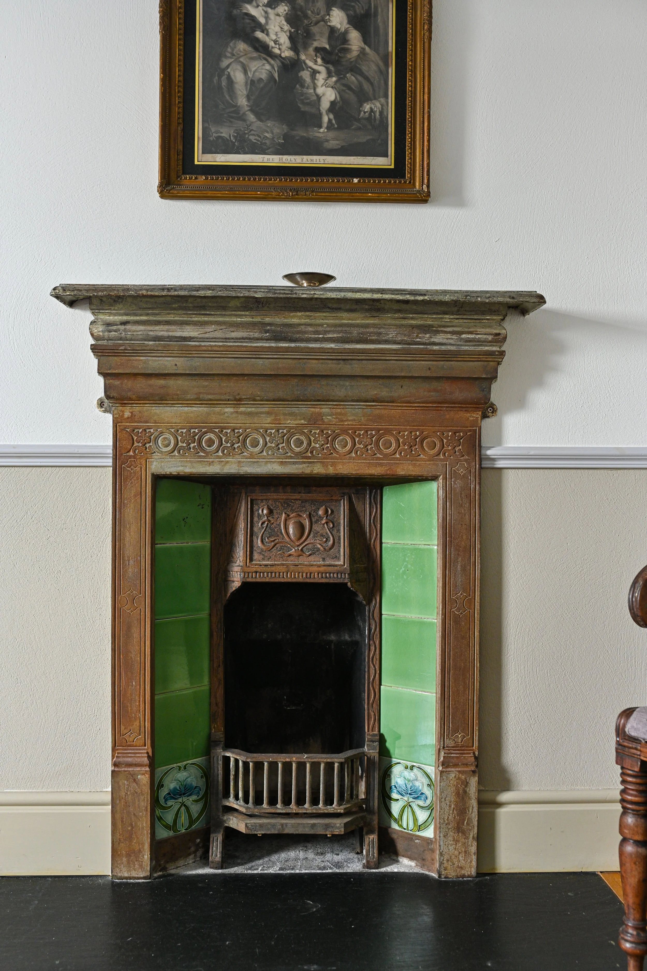 An antique fireplace with green tile accents and a carved wooden mantel, with a framed religious artwork hanging above it.