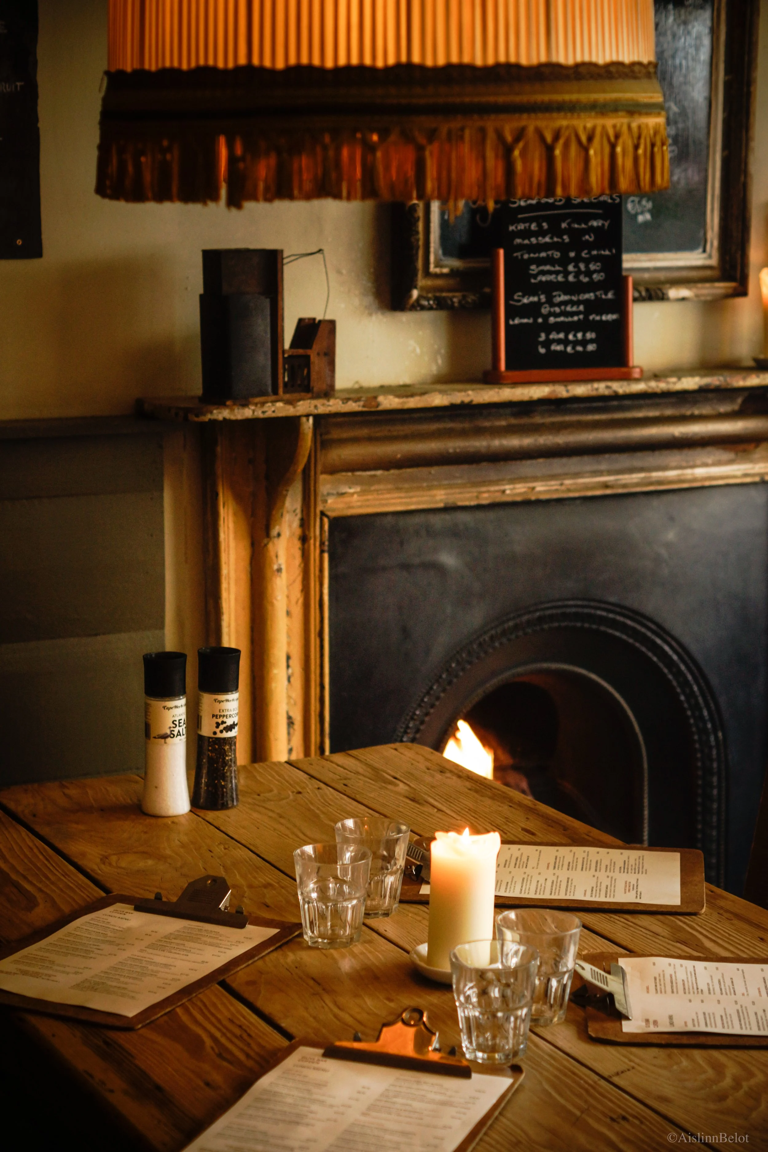 A wooden table set with glasses, menus, and a lit candle in a cozy restaurant with a fireplace, a chalkboard menu, and a fringed lampshade.