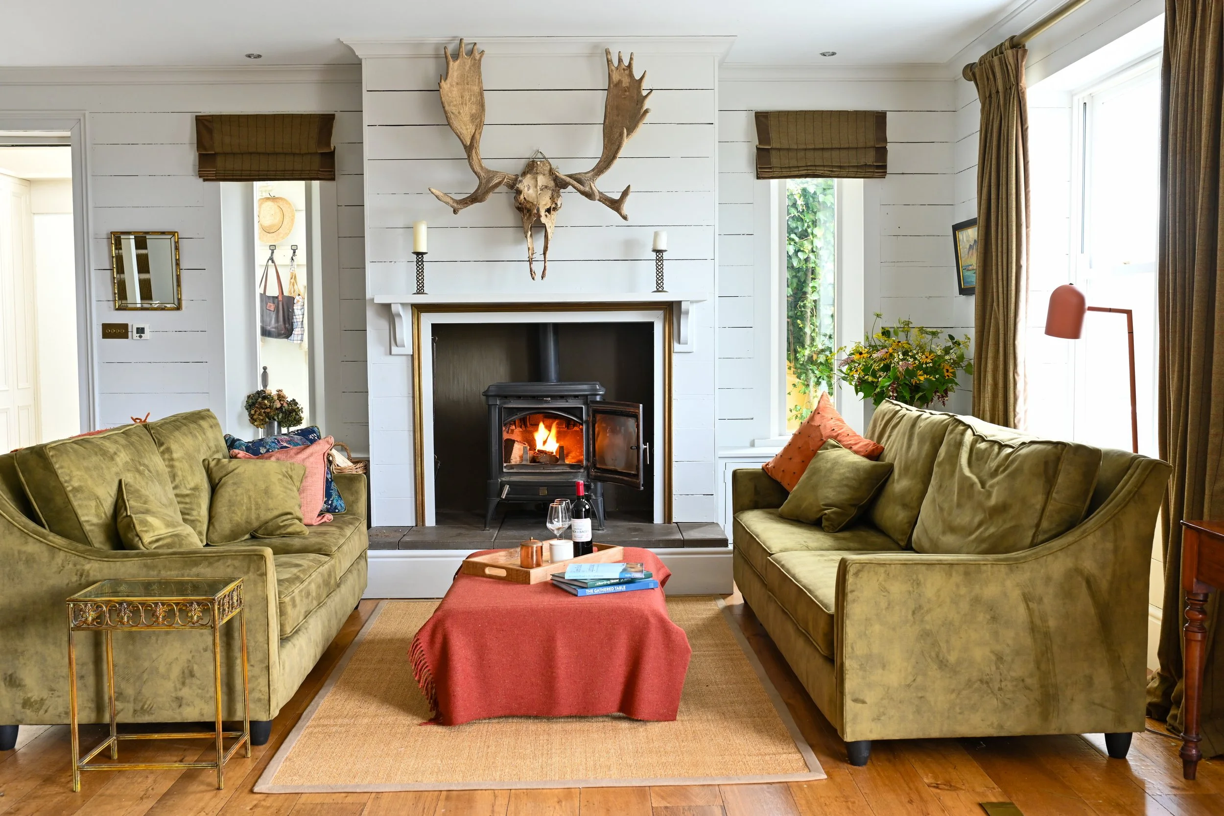 Living room with a fireplace, mounted moose skull with antlers above, two green sofas with pillows, a red tablecloth on a coffee table, and large windows with curtains.