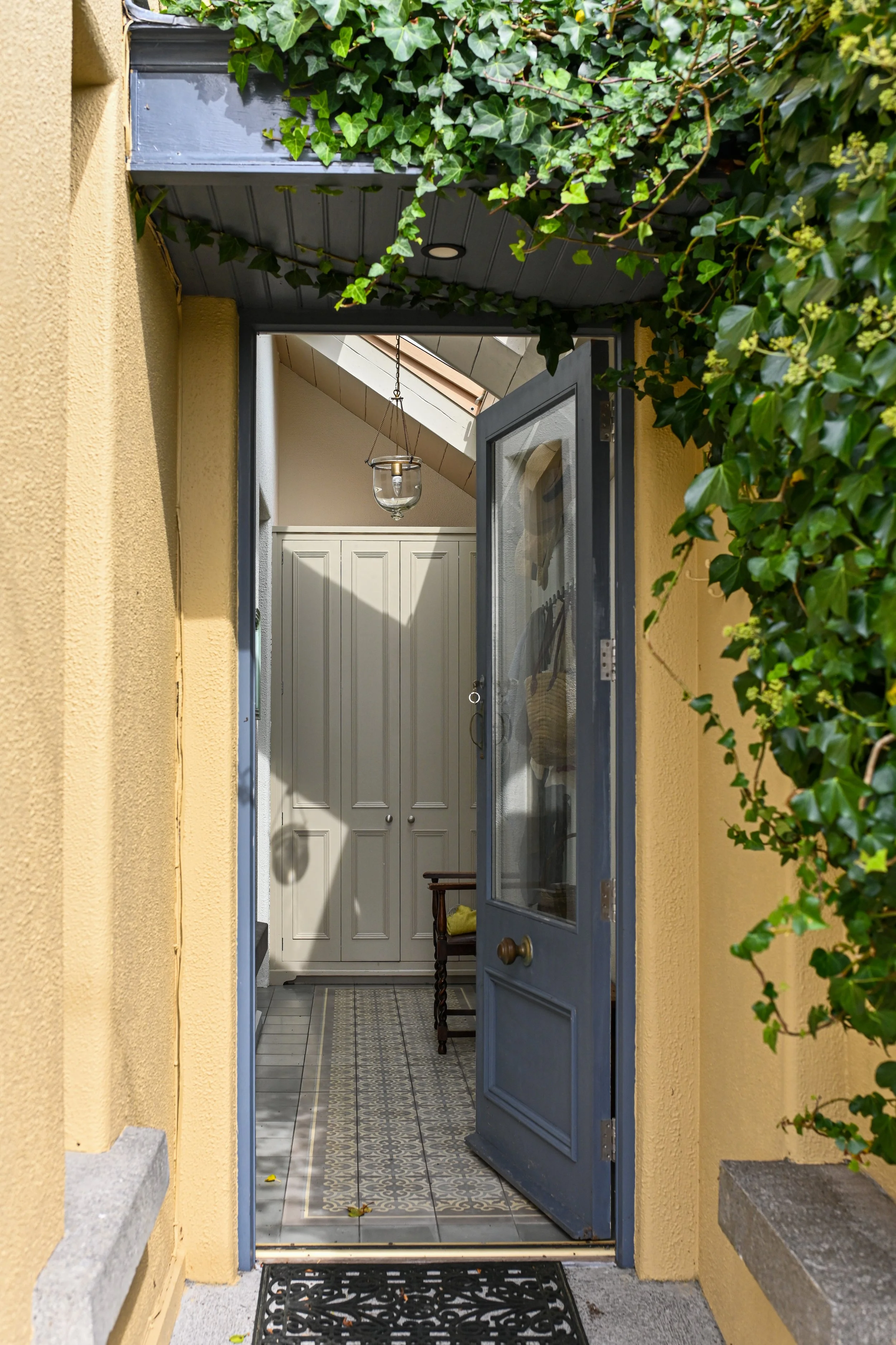 Open blue front door of a house, revealing a small entryway with patterned tile floor, beige walls, a white cabinet, and a small wooden bench. Green ivy surrounds the doorframe.