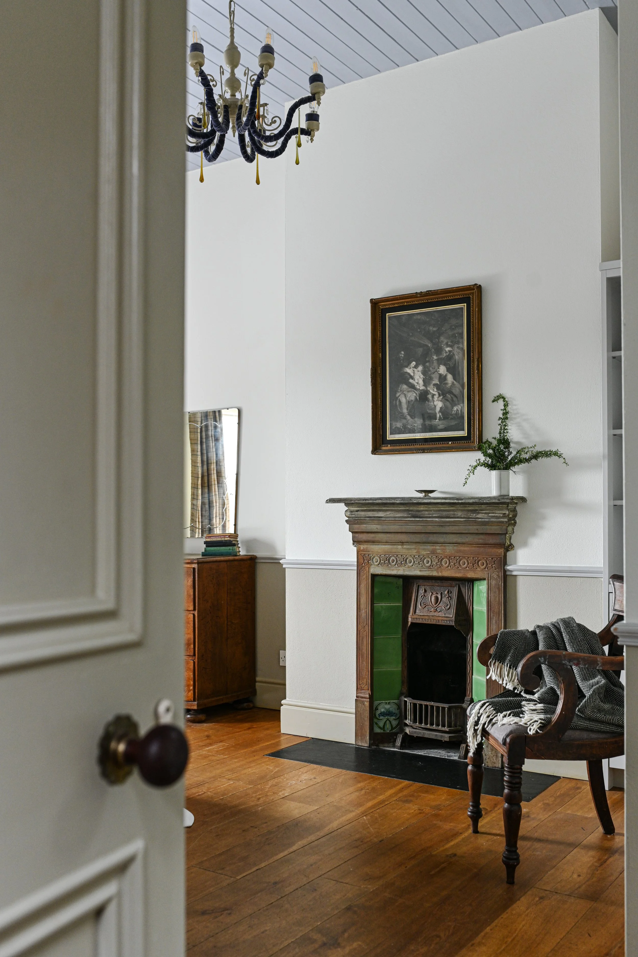 Photo of a living room with a vintage fireplace, framed artwork above, and a wooden armchair with a blanket on it. A chandelier hangs from the ceiling, and a small mirror with books on a cabinet is visible, through an open door.