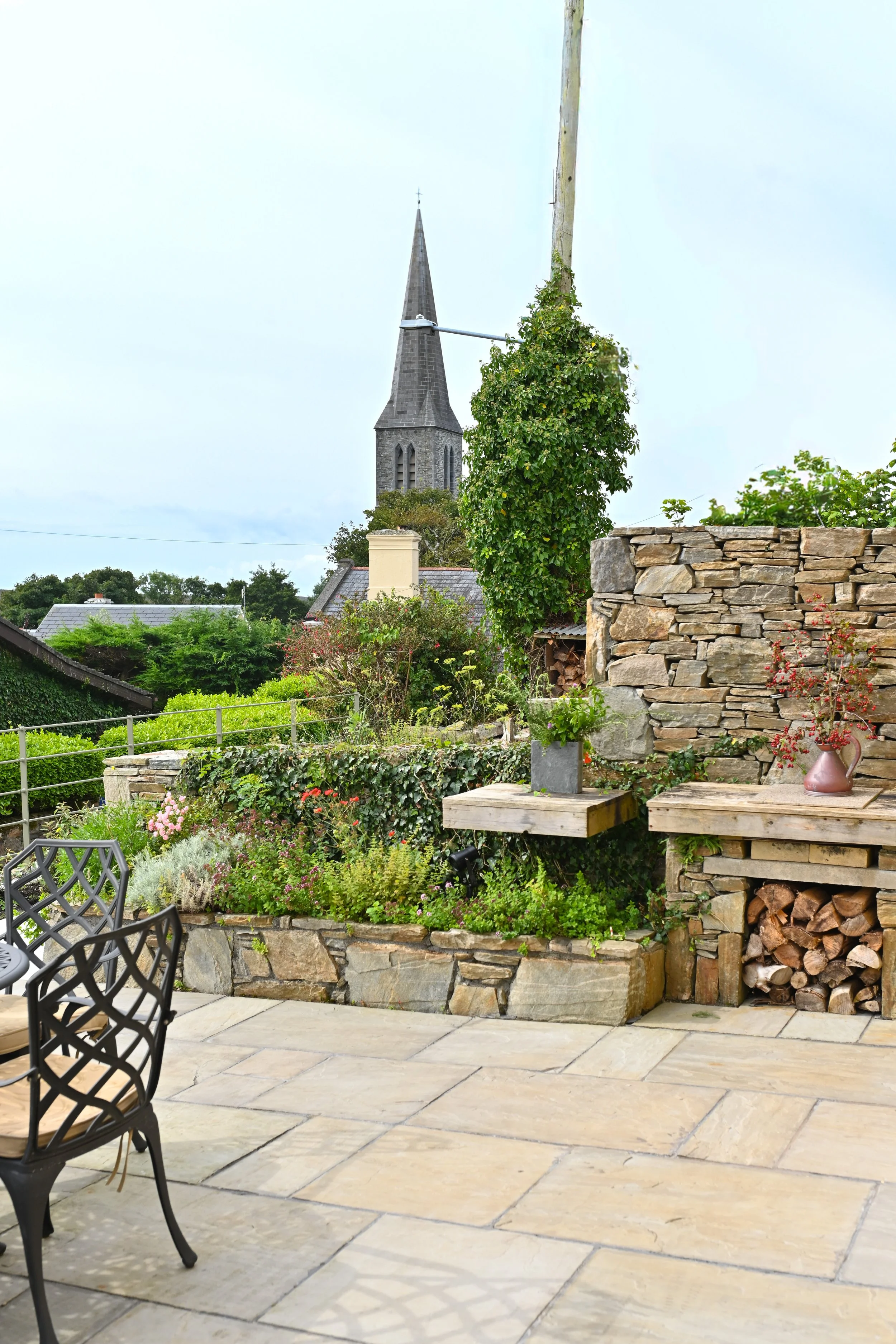 A stone patio with black chairs, a garden bed with flowers and green plants, a stone wall with stacked firewood, a vineyard, and a tall church steeple in the background.