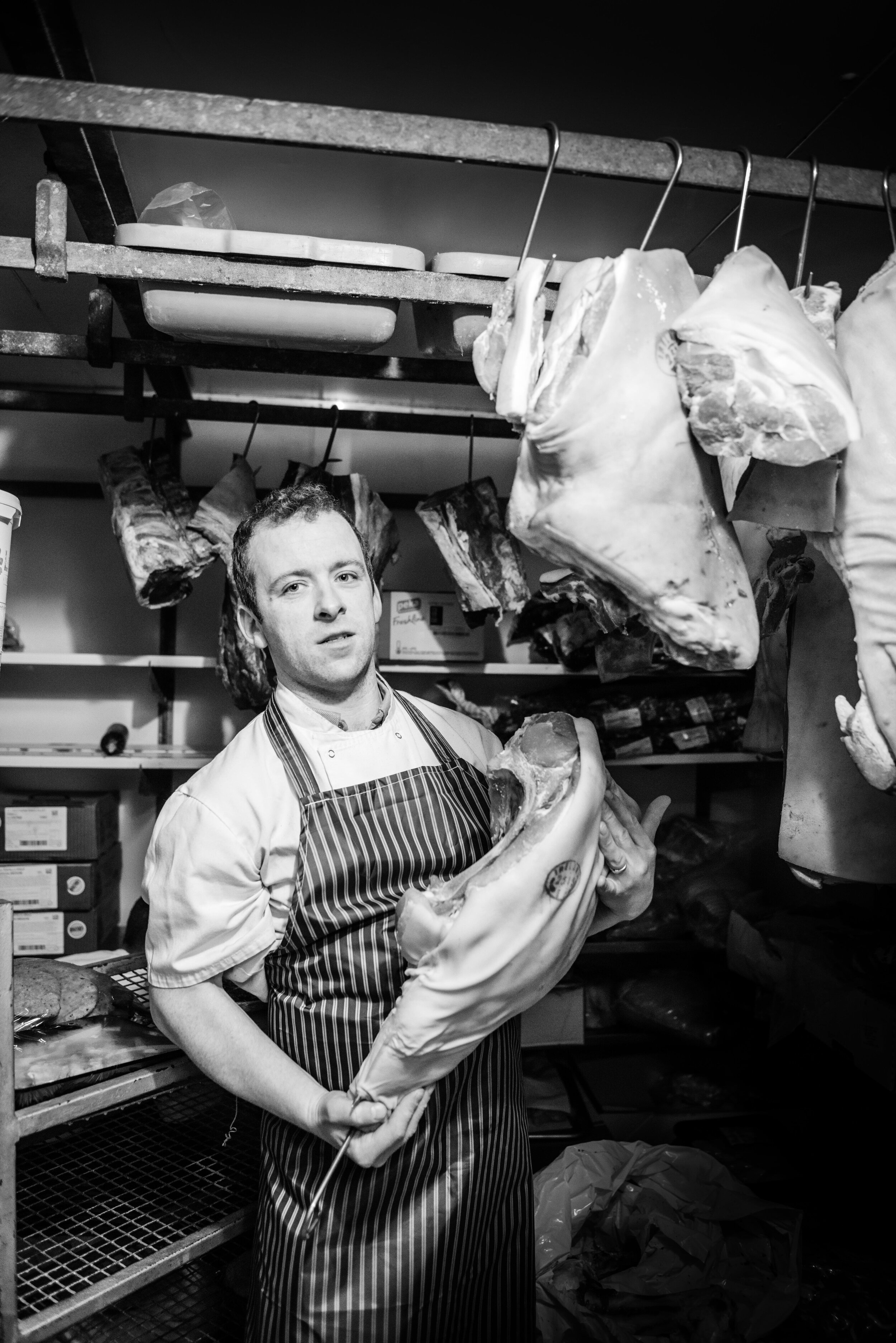 Butcher holding a large cut of meat in a meat locker, surrounded by hanging meat cuts.