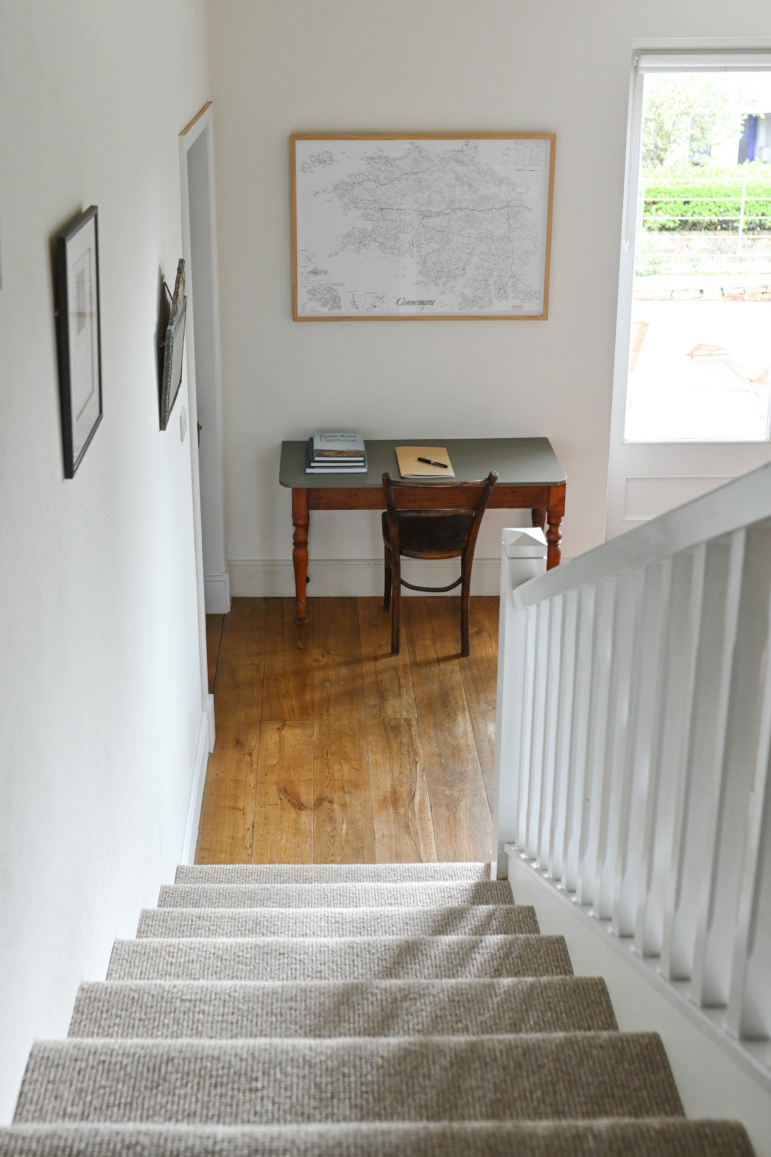 A staircase leading down to a small landing area with a desk, a chair, and framed maps on the wall. Natural light coming in through a large window.