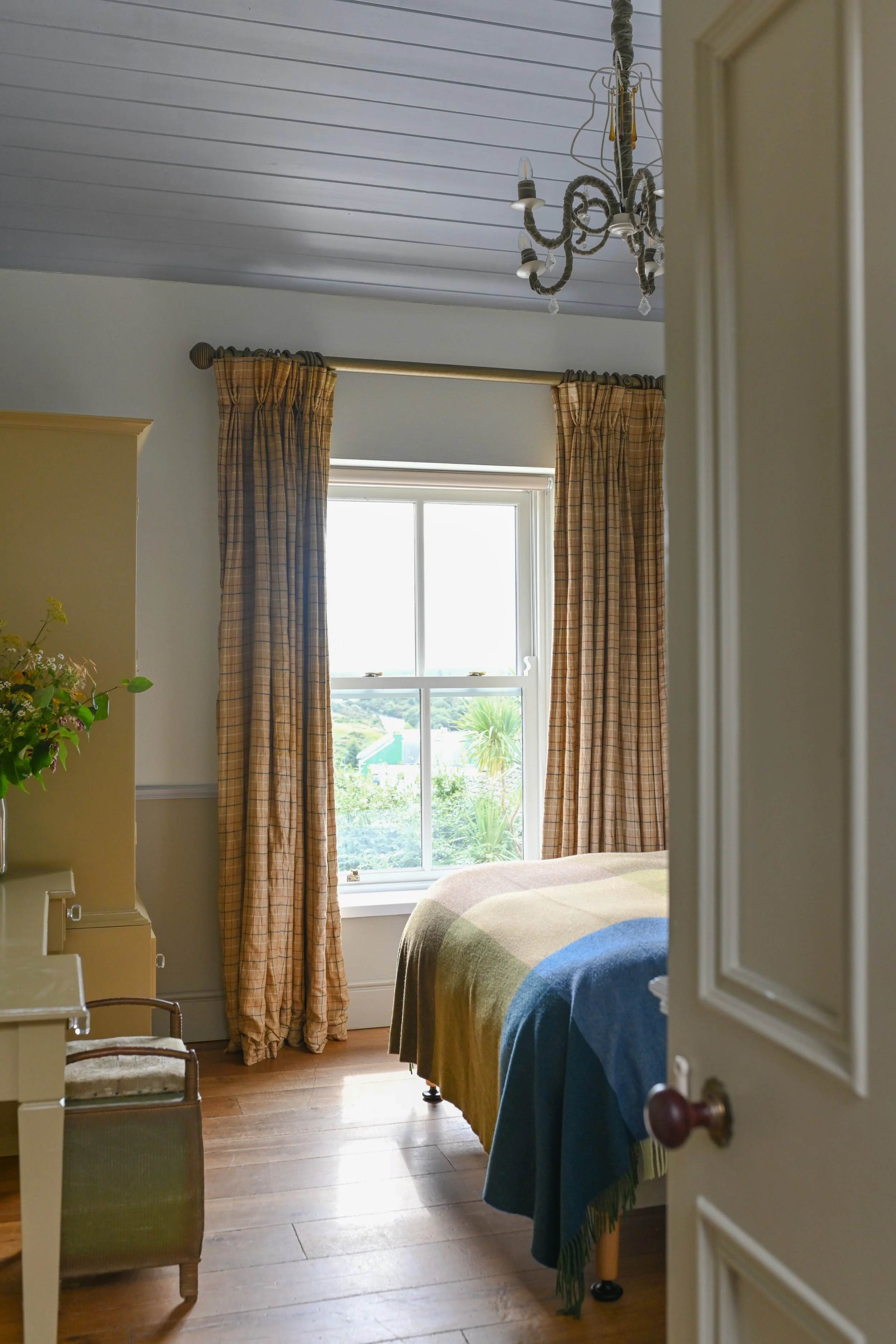 View of a cozy bedroom with a large window, yellow plaid curtains, a bed with a colorful blanket, a small chair, and a plant on a side table. There's a chandelier hanging from the ceiling.
