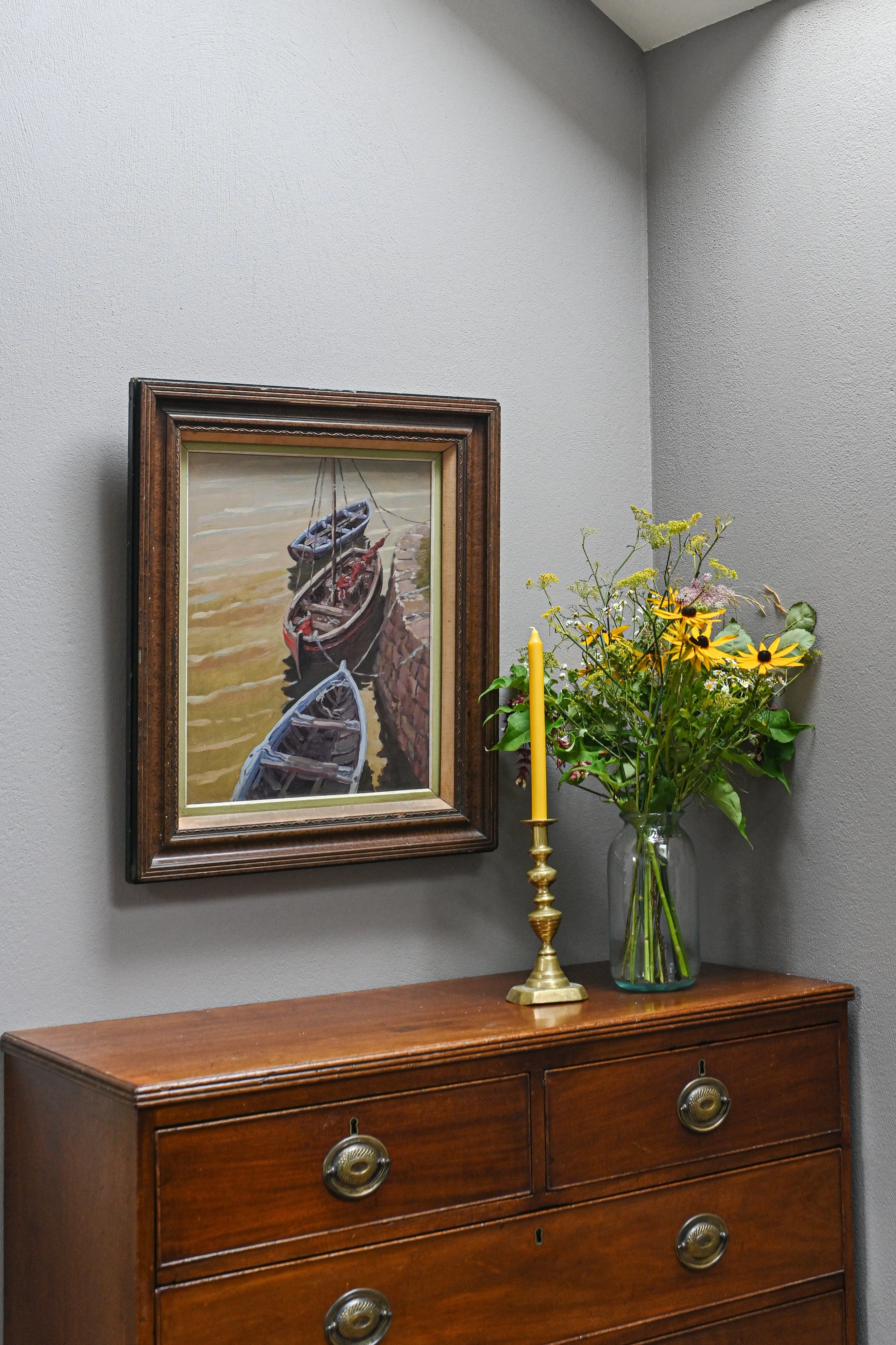A wooden dresser with brass handles. On top, there is a yellow candle holder with a tall yellow candle and a glass vase with a bouquet of yellow and white flowers. A framed painting of boats docked along a shoreline hangs on the gray wall behind.