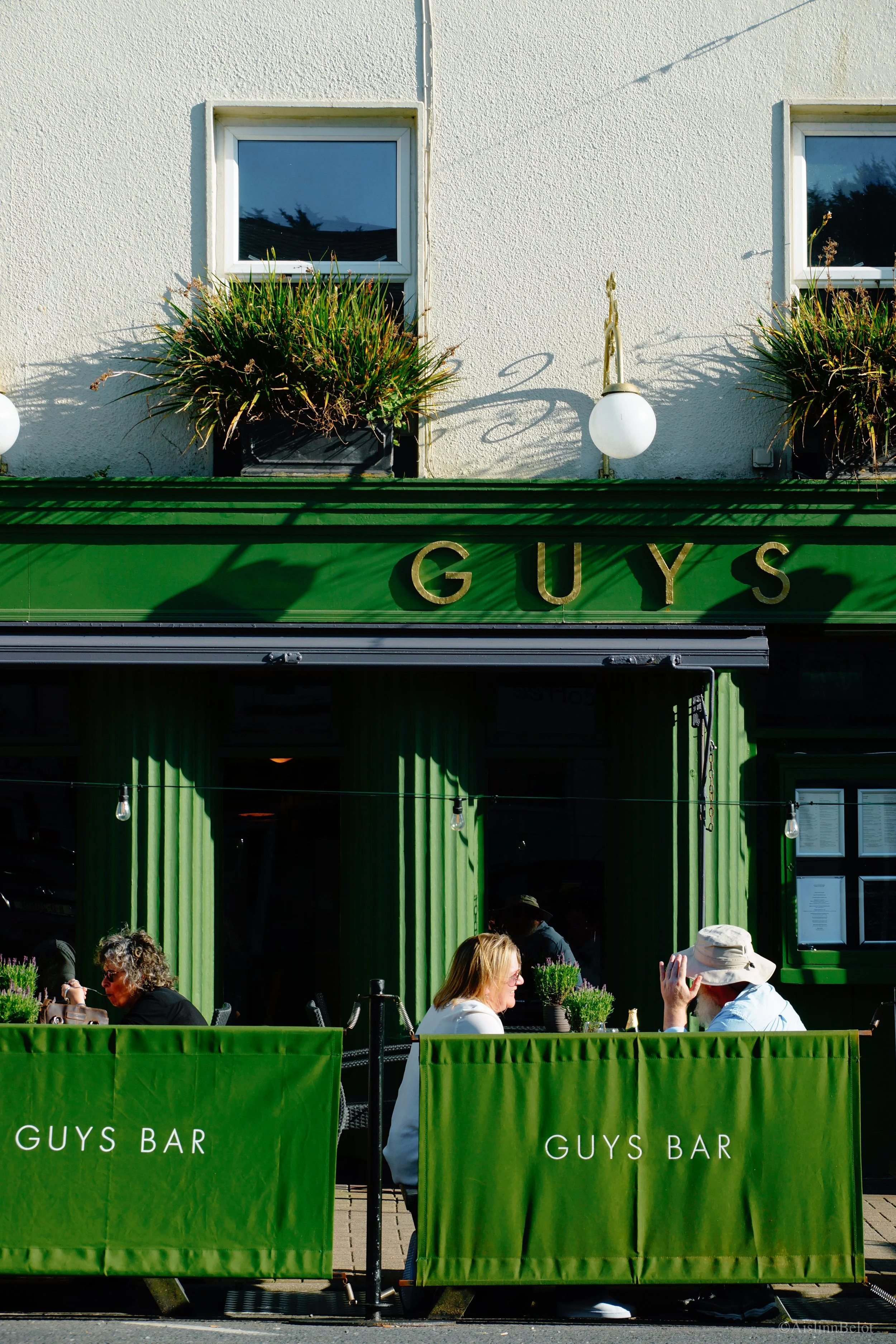 Front view of a green bar called 'Guys Bar', with outdoor seating where three people are sitting and talking.