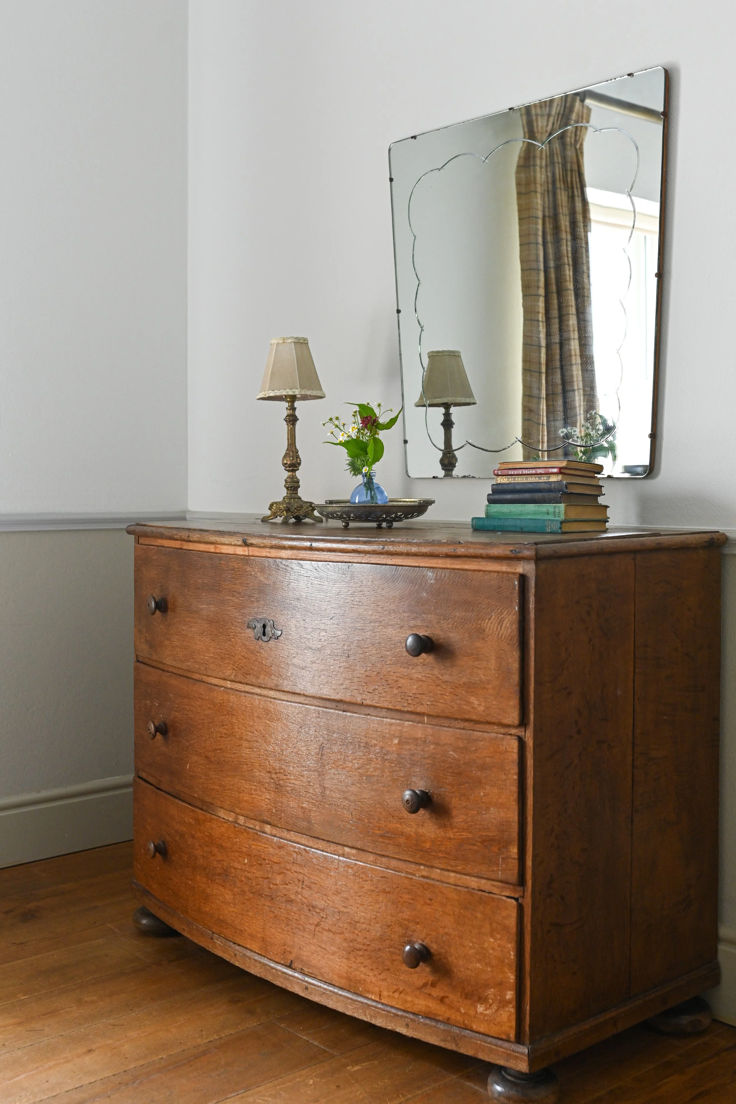 Vintage wooden dresser with a mirror above it, decorated with a small table lamp, a vase with flowers, and a stack of books, with a window and plaid curtain reflected in the mirror.