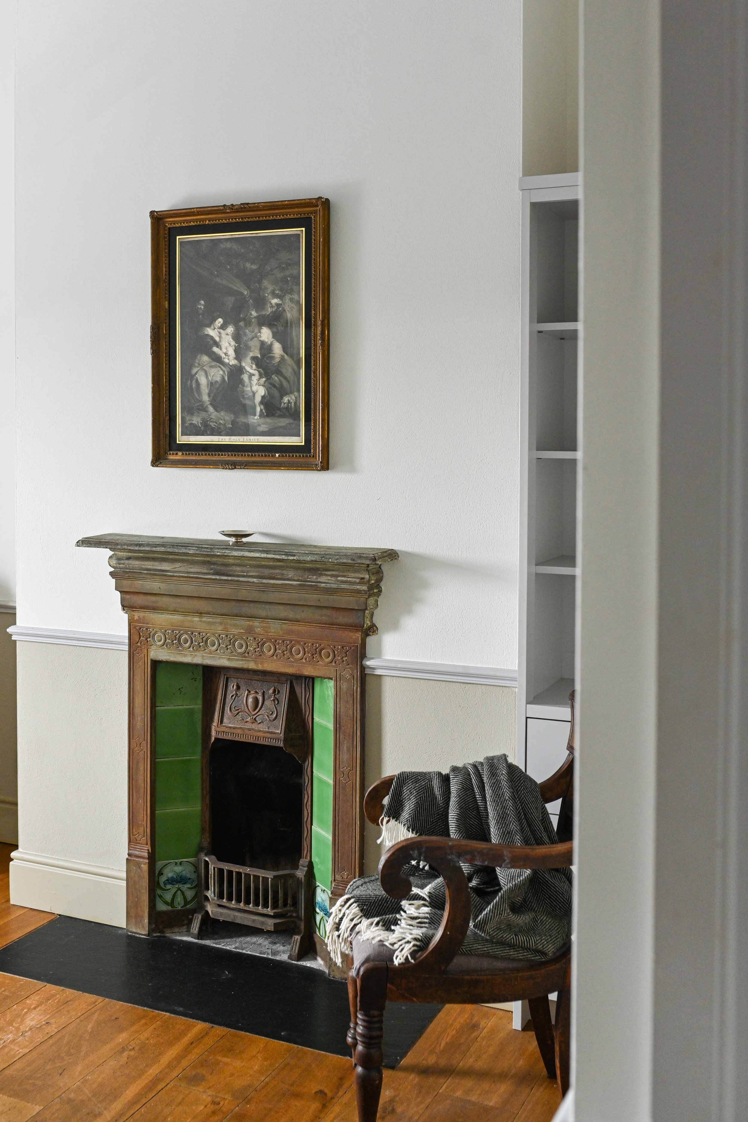 Living room with a vintage fireplace with green tiles, a wooden chair with a black and white blanket, a framed painting above the fireplace, and a white built-in bookshelf on the right.