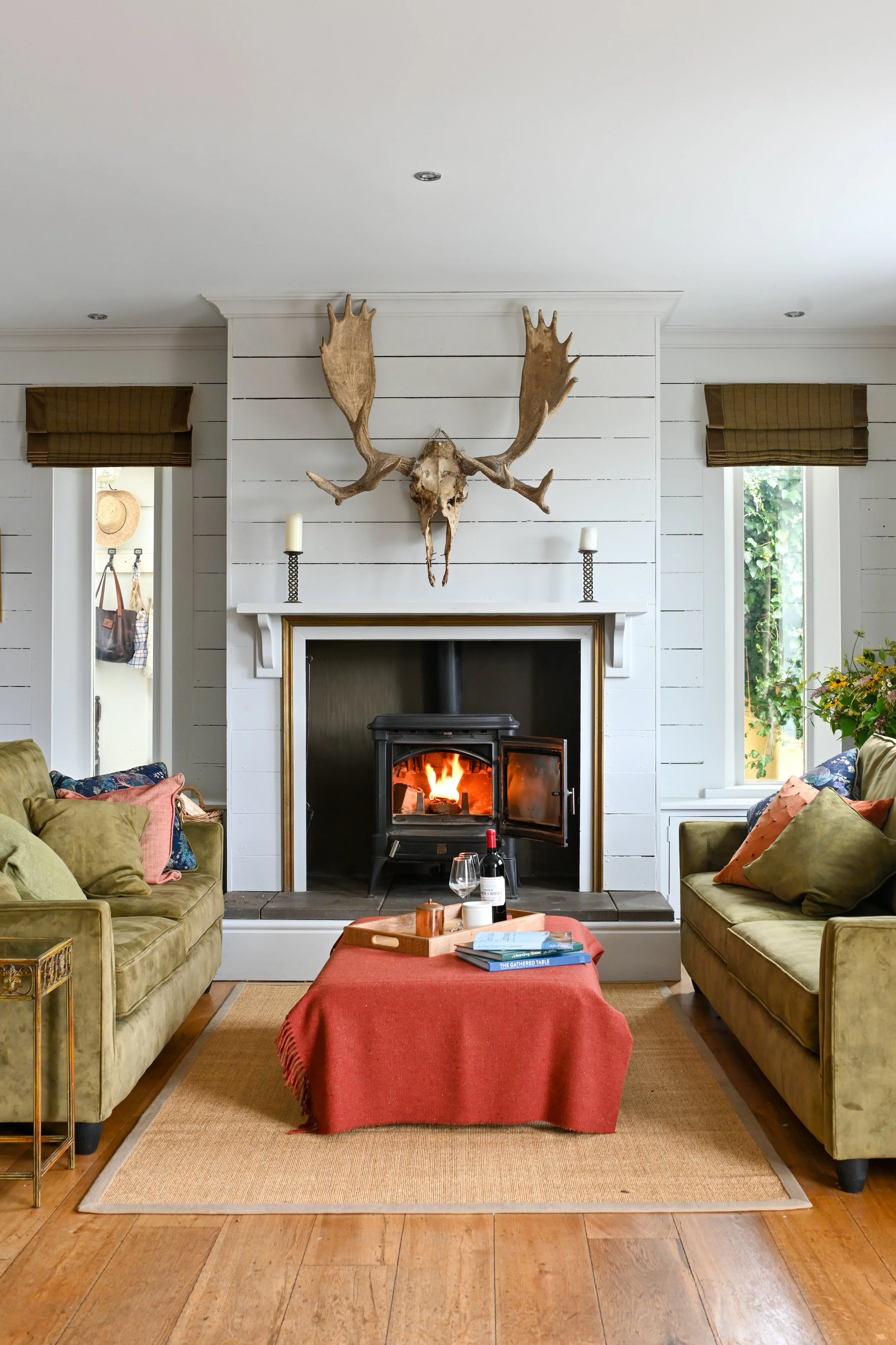 Living room with a fireplace, moose antlers and skull mounted above, green sofas with colorful pillows, a red-covered coffee table with a wine bottle and books, and windows on both sides of the fireplace.
