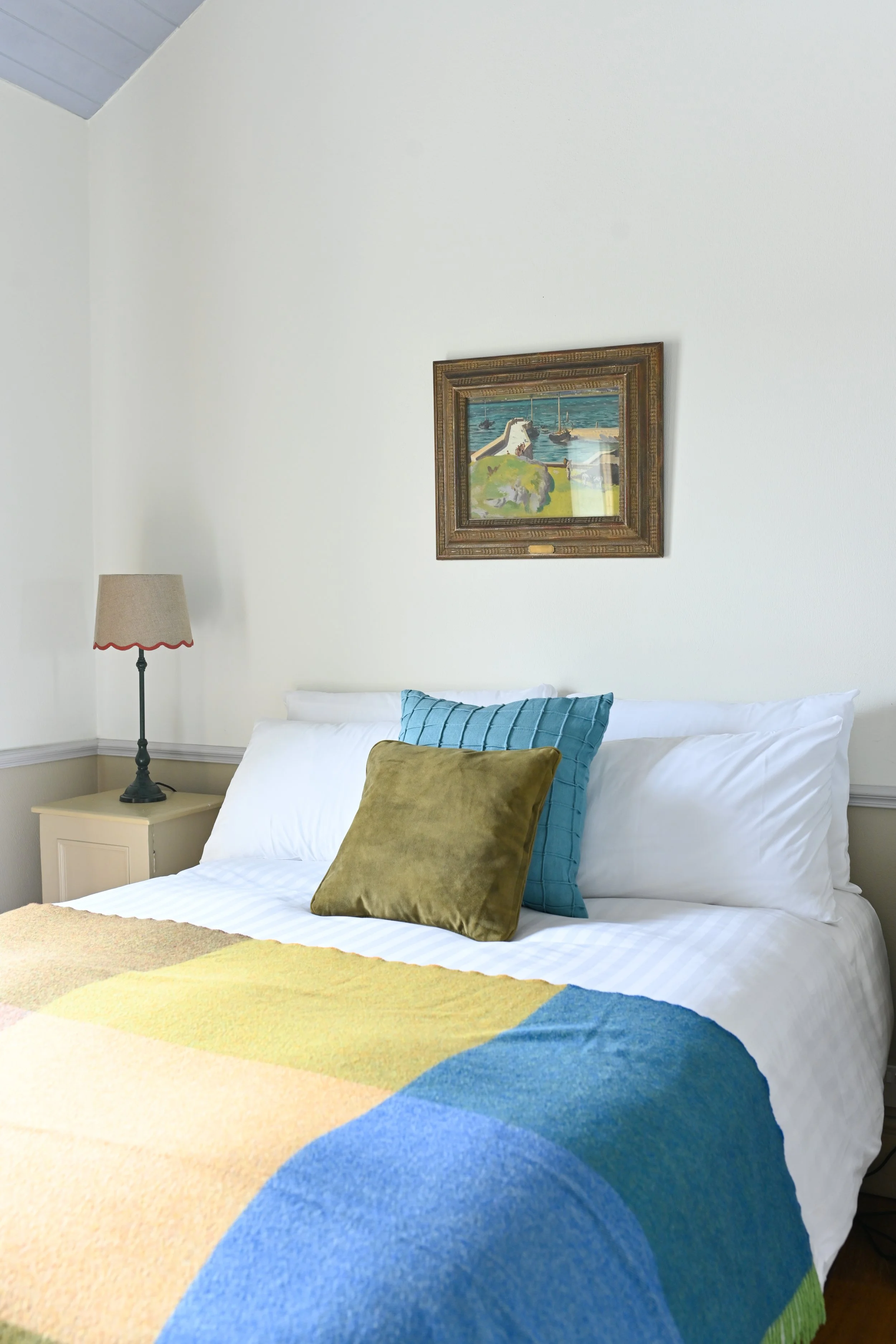 A neatly made bed with white sheets, two white pillows, a blue pillow, and a green pillow, covered by a colorful quilt. To the left, there's a beige nightstand with a lamp. A framed painting of a coastal scene hangs above the bed. The wall is white,