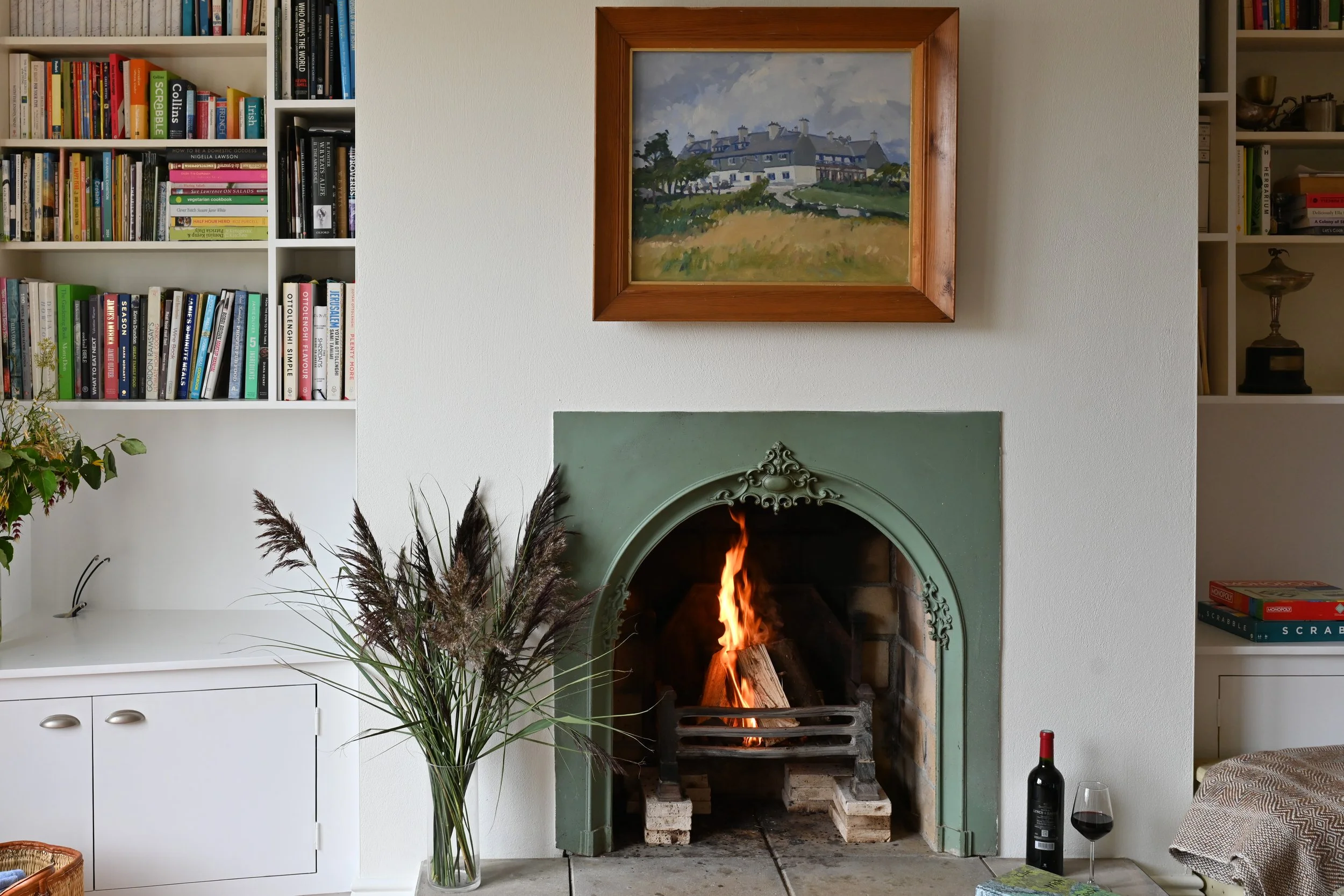 Living room with a fireplace burning, flanked by framed paintings, white bookshelf filled with books on both sides, a large vase of tall grasses on a white cabinet, a bottle of wine and a glass of red wine on the floor, and part of a couch with a pat